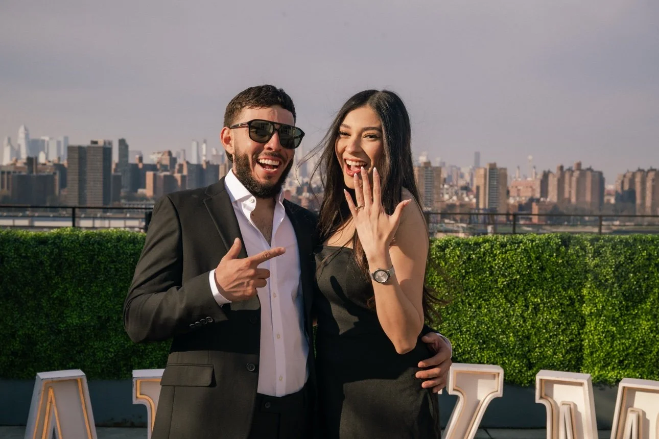 A man and woman standing on a rooftop with a city skyline in the background. The man is wearing a suit and sunglasses, pointing at the woman. The woman is wearing a black dress, smiling, and showing her ring. They are posing together in a joyful mome