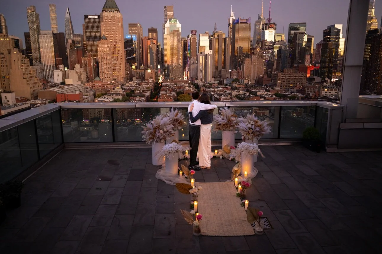 A couple stands on a rooftop with a view of a city skyline at sunset, surrounded by floral arrangements and candles, embracing each other during their wedding ceremony.