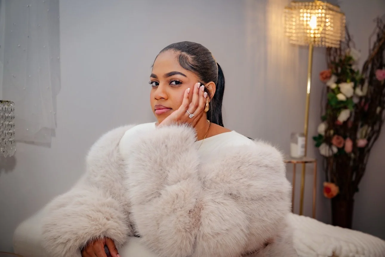 A woman with dark hair tied back, posing indoors with her hand on her face, wearing a white fur coat. The background features a modern lamp, a small table with a bottle, and flower arrangements.