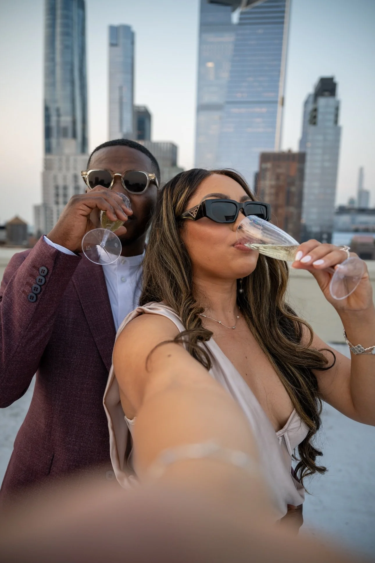 A man and woman in stylish clothing, wearing sunglasses, celebrating and taking a selfie on a rooftop with a city skyline in the background while drinking champagne.