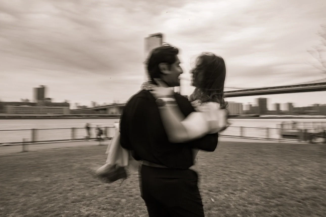 A man and woman embracing outdoors near a river, with city buildings and a bridge in the background, in black and white.
