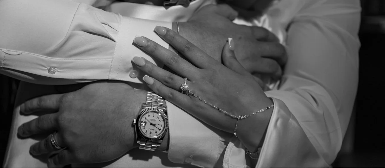 Close-up of a couple's hands, showcasing wedding rings, a silver watch, and a bracelet, with the woman’s hand resting on the man's arm, on a formal occasion.