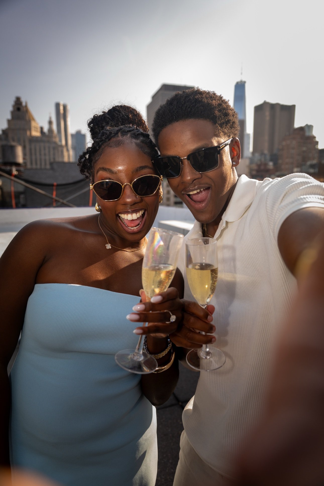 A smiling woman and man wearing sunglasses, holding champagne glasses, celebrating on a rooftop with city buildings, including One World Trade Center, in the background.
