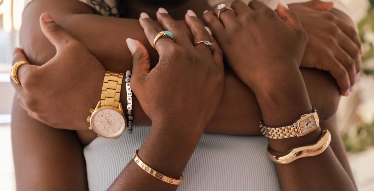 Multiple people with dark skin tones hugging each other, showing their arms and wrists adorned with various gold and silver watches, bangles, and rings.