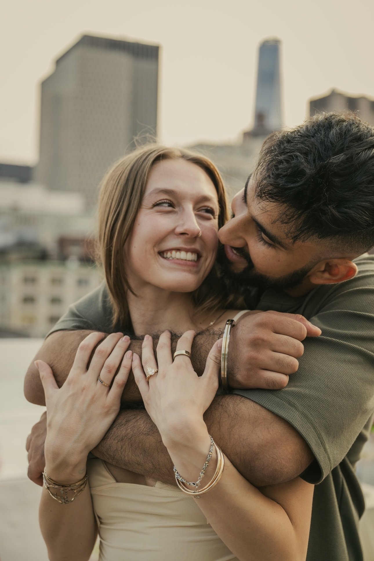 A smiling young woman with light brown hair embraces a man with dark hair and a beard on a rooftop with city buildings in the background.