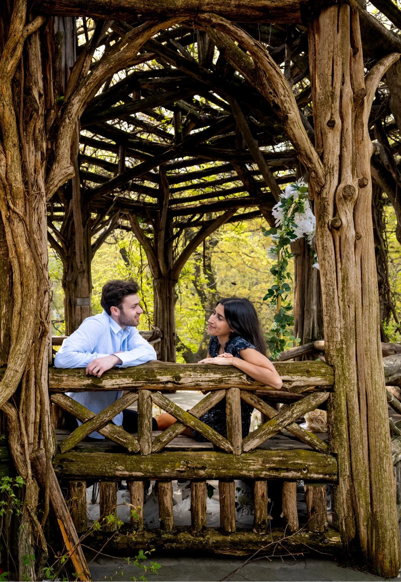 A man and woman sitting on a wooden balcony inside a rustic, tree-like wooden structure, looking at each other, surrounded by greenery.