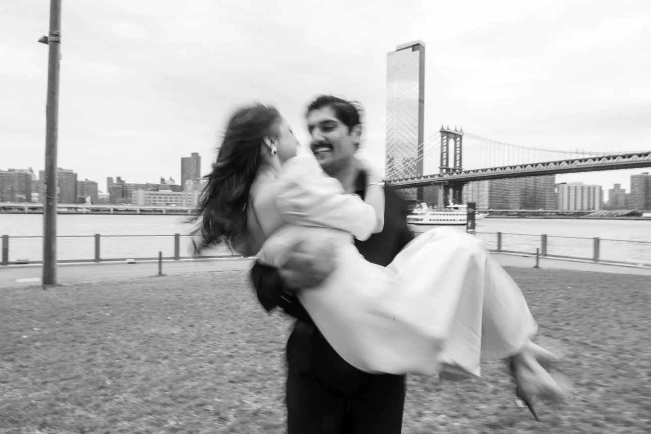 A man is holding a woman in his arms by the waterfront, with the Brooklyn Bridge and Manhattan skyline in the background, in black and white.