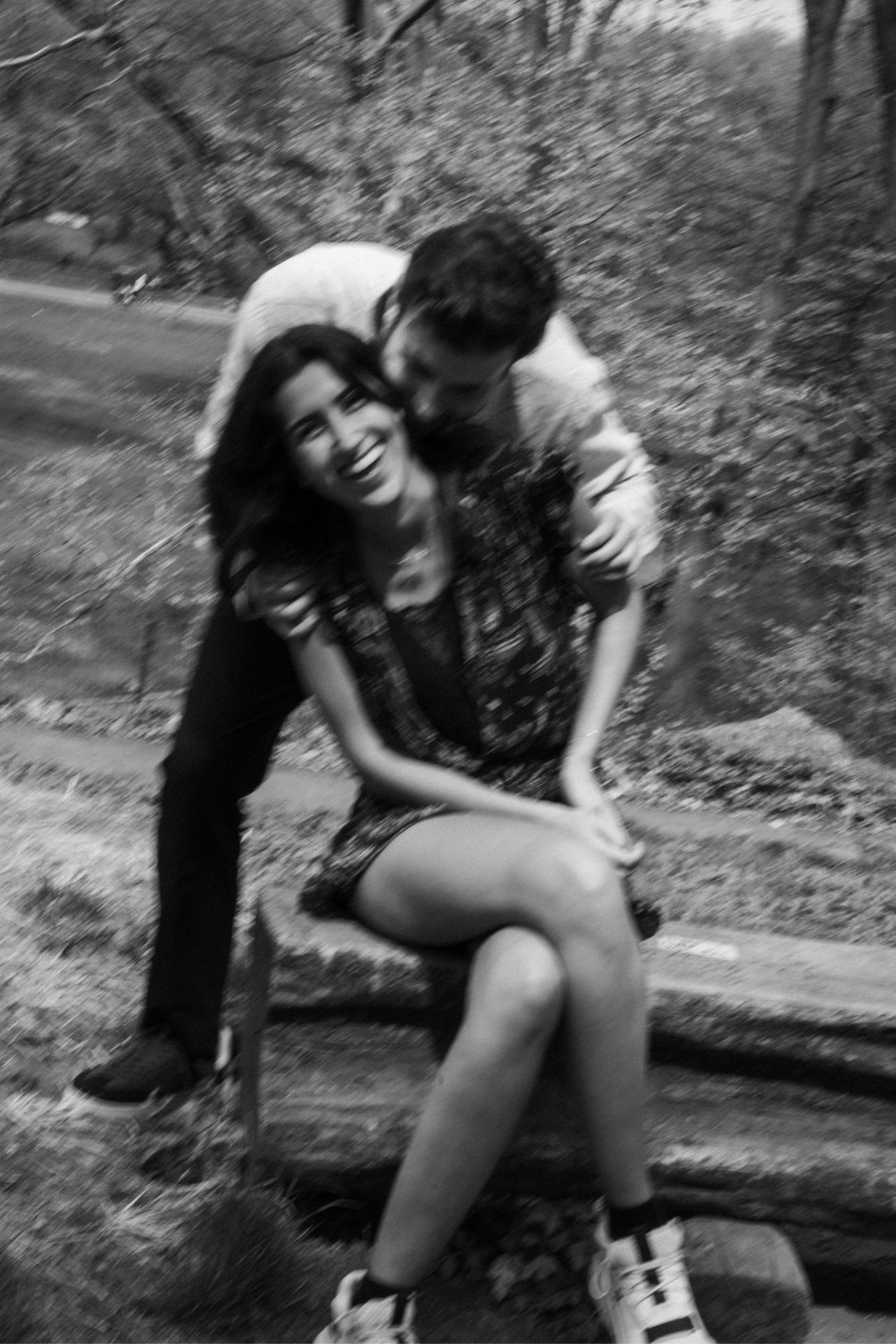 A black and white photo of a young woman with dark hair smiling and a man kissing her cheek, with the woman seated on a scooter or motorcycle and the man leaning in close behind her. They are outdoors near a wooded area.
