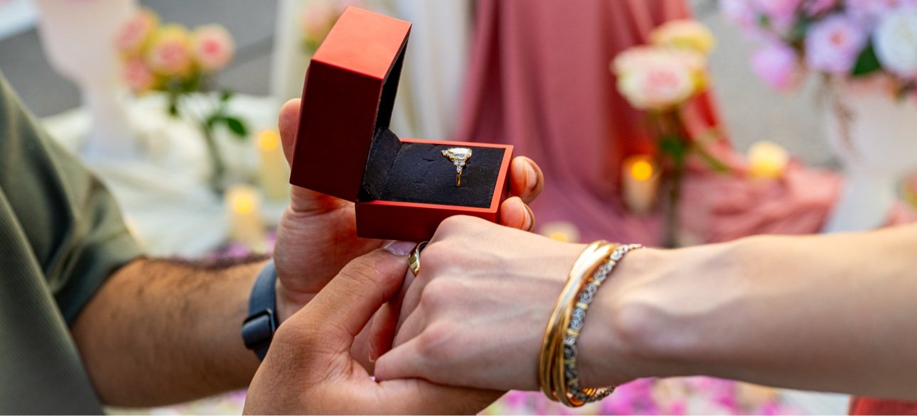 A couple is exchanging wedding rings; the man is holding an open red ring box with a diamond ring inside, while the woman is reaching to place the ring on the man's finger, with her hand adorned with gold jewelry. The background features pink and whi