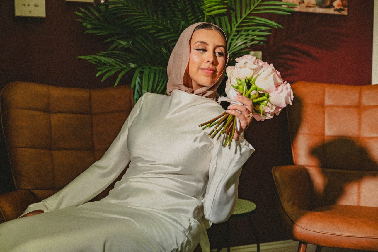 A woman in a white satin dress and beige headscarf sitting on a brown chair, holding a bouquet of pink roses, with green plants and a dark wall behind her.