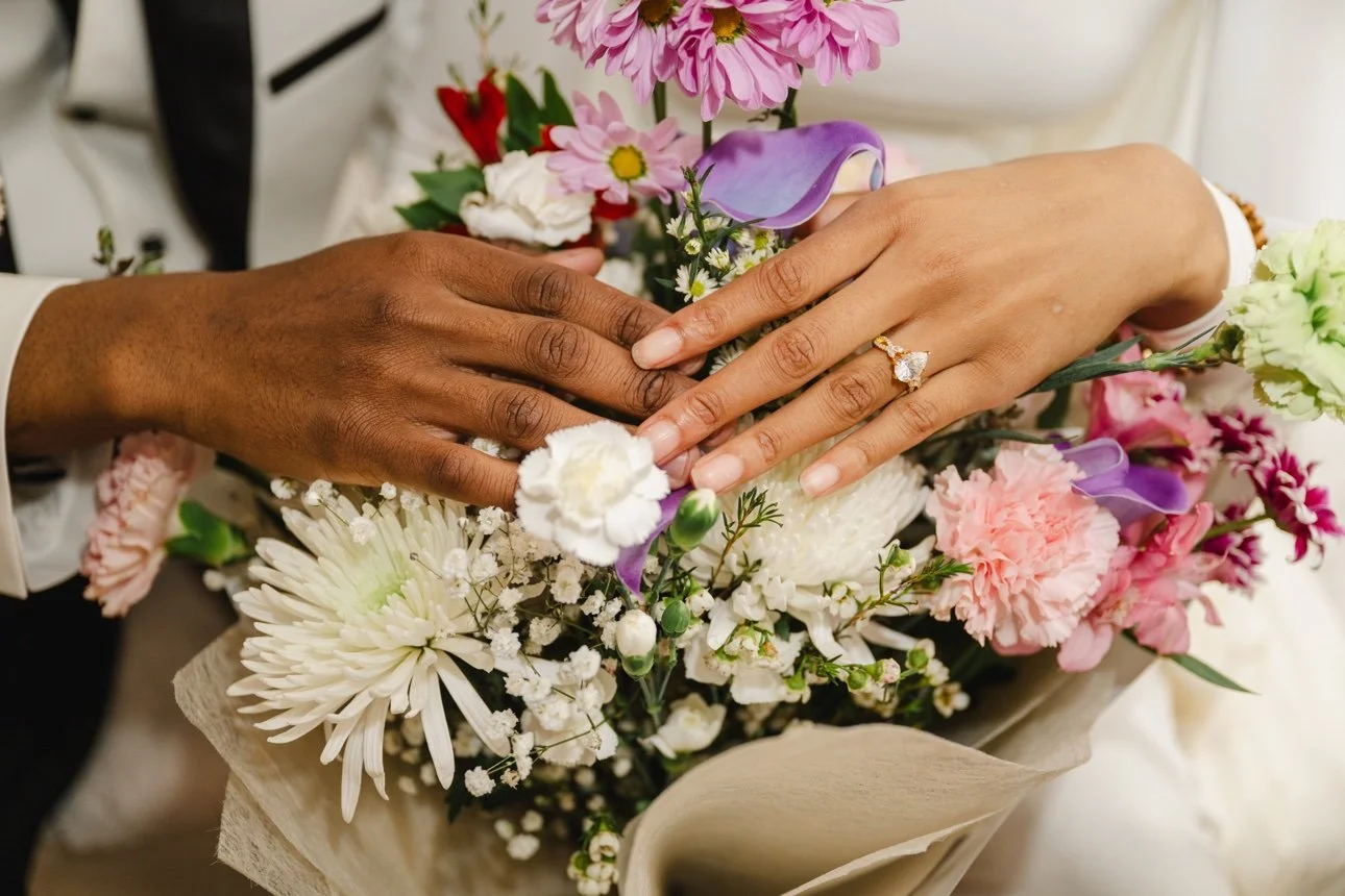 Two hands with wedding rings resting on a bouquet of assorted pink, white, and purple flowers.