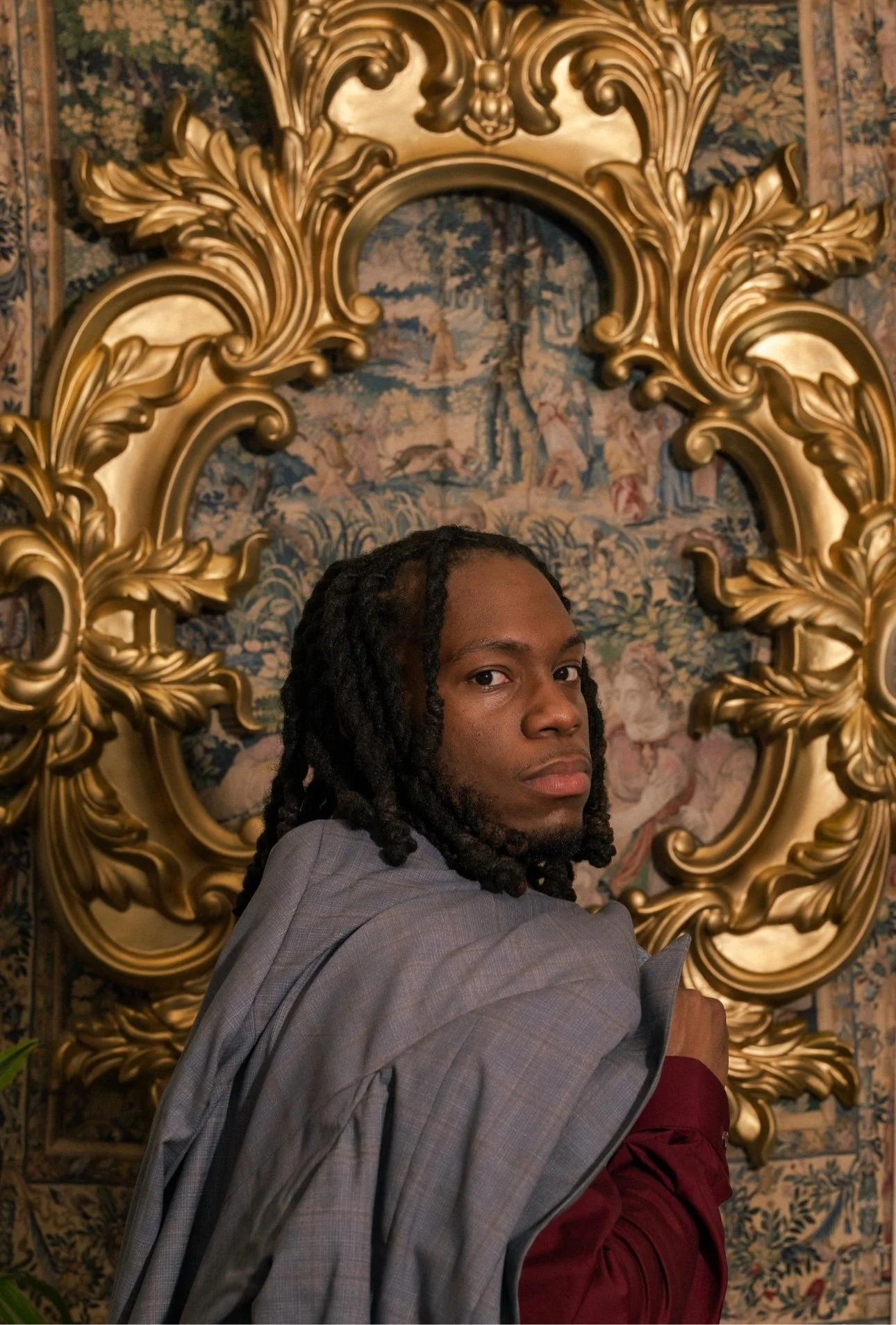 A man with dreadlocks and a gray suit looking over his shoulder, seated in front of an ornate gold decorative frame and a tapestry background.