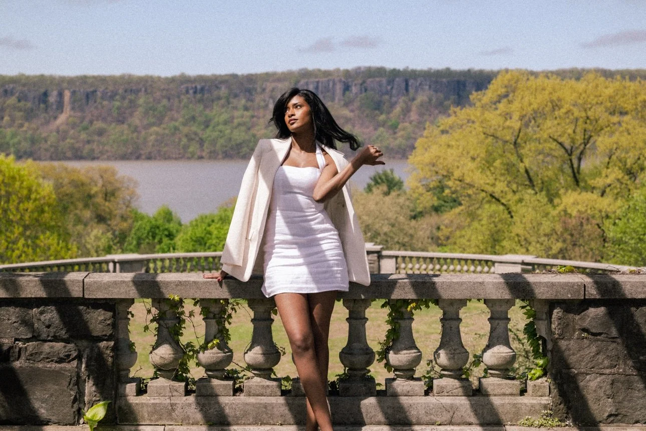 A woman in a white dress and blazer leaning against a stone railing outdoors, with trees, a lake, and hills in the background on a sunny day.