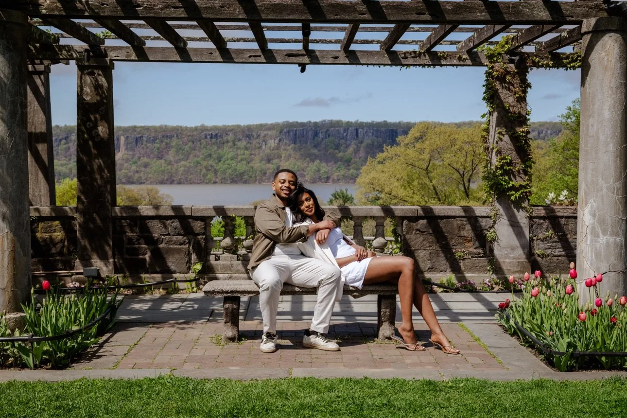 A couple sitting close together on a stone bench outdoors under a wooden pergola, smiling and embracing, with a scenic view of a lake, trees, and cliffs in the background, and pink tulips in flower beds in front.