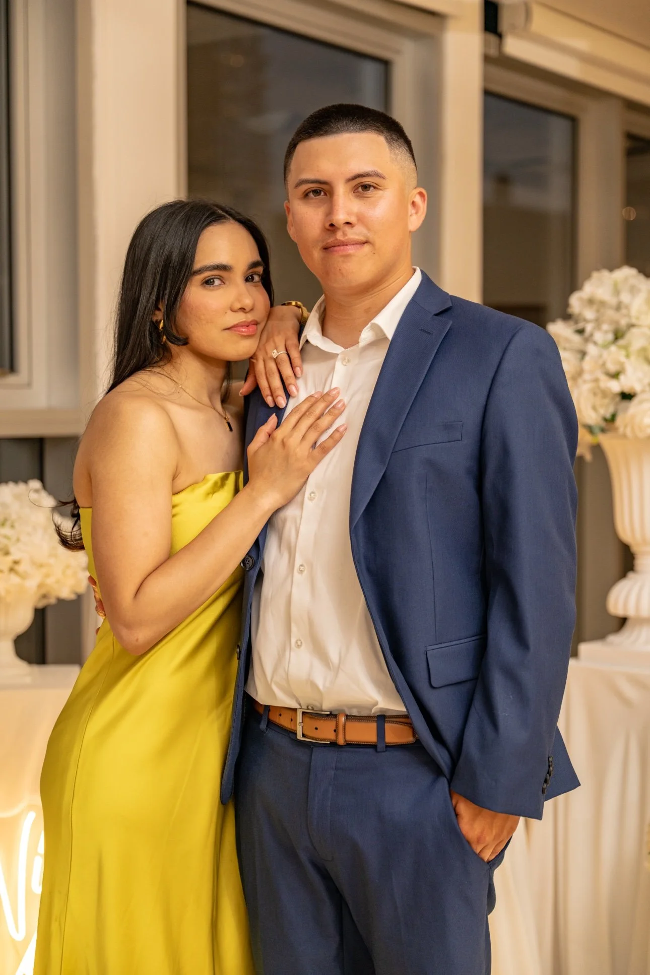 A woman in a yellow dress and a man in a blue suit posing together indoors with floral arrangements in the background.