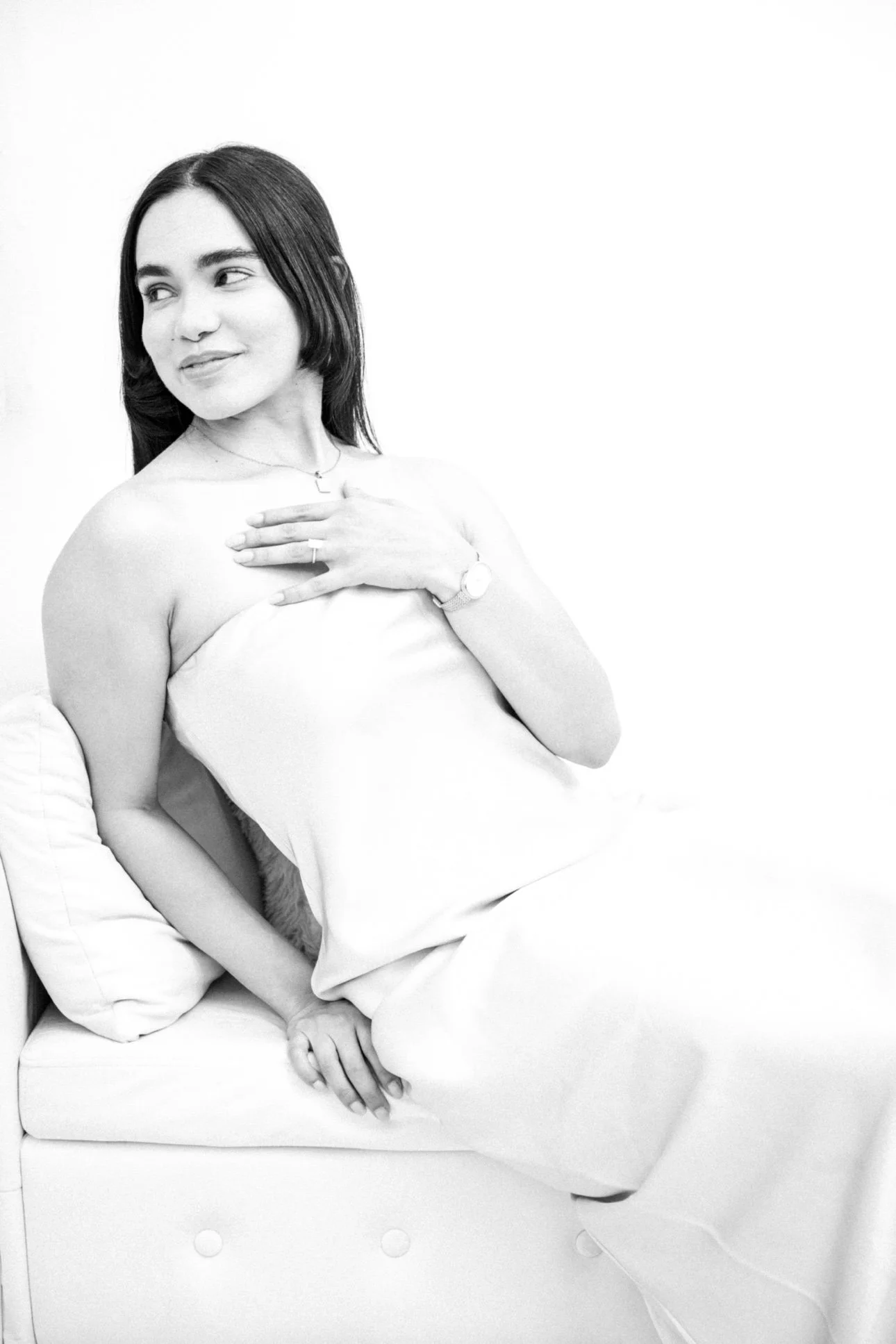 Black and white photo of a woman with dark hair sitting on a bed, looking to her left, smiling softly, with one hand on her chest and the other resting on her lap, surrounded by white bedding.