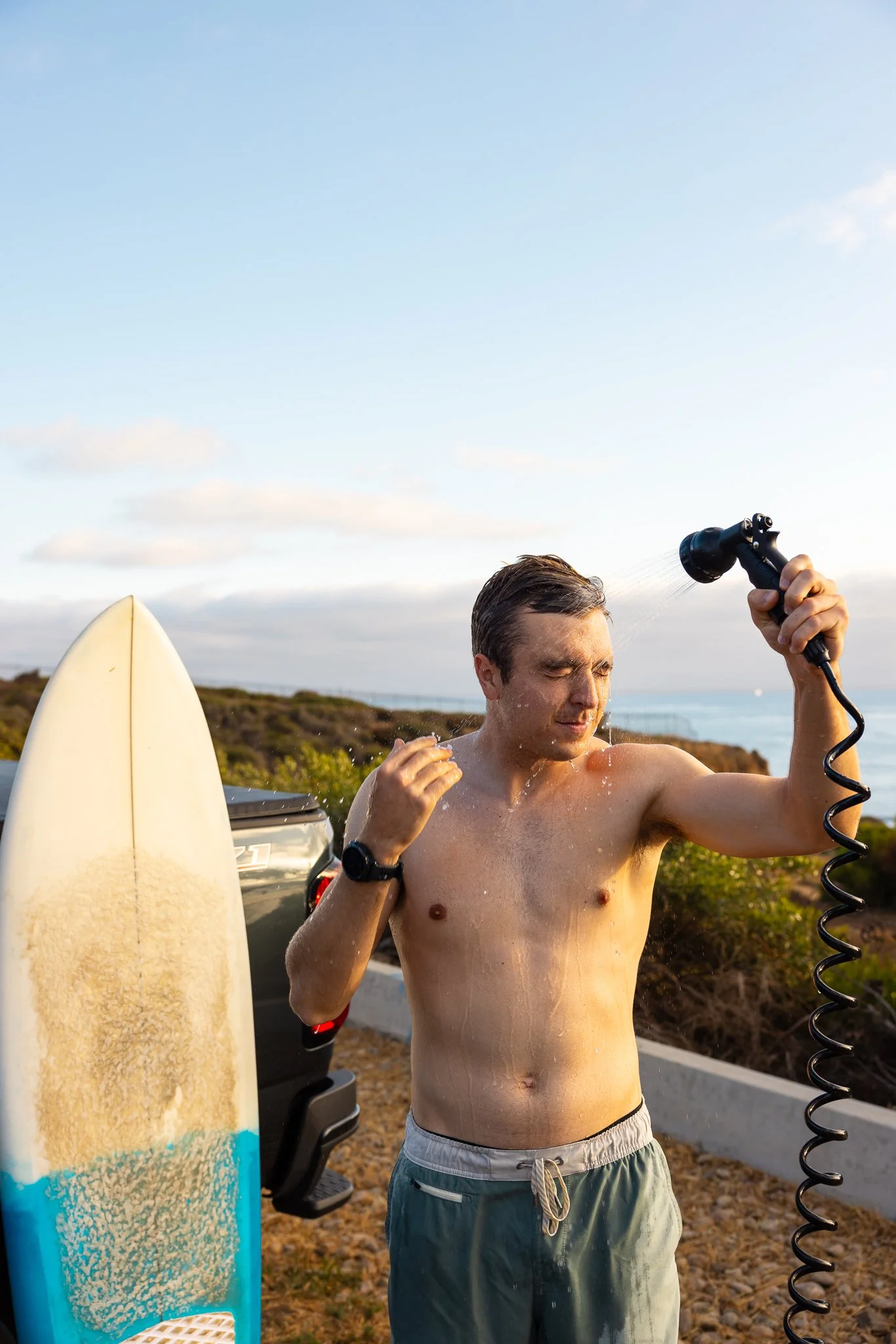 A man rinsing off with a garden hose near a surfboard at the beach during sunset.