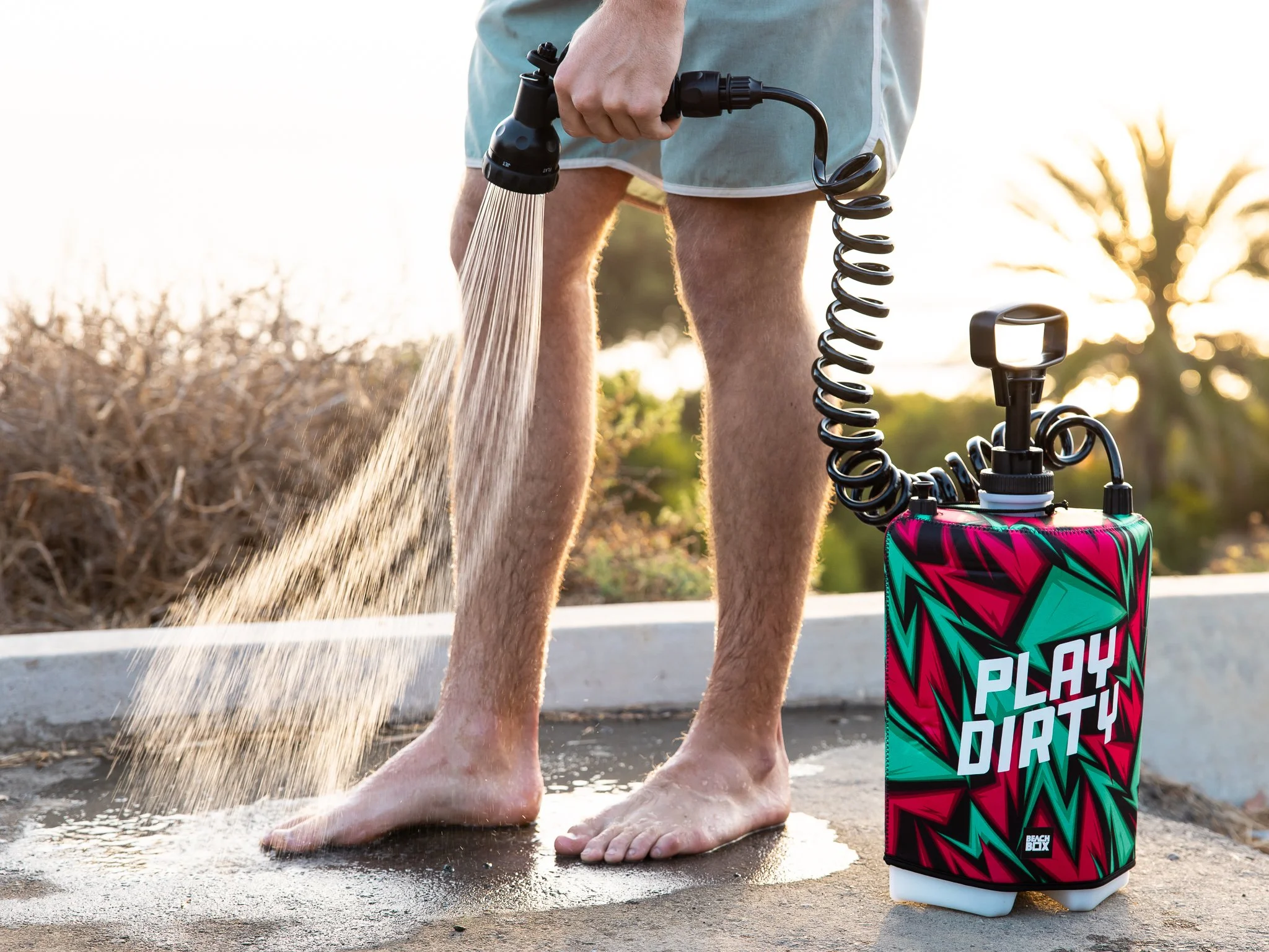 Person standing barefoot outdoors, watering their feet with a portable spray tank labeled "Play Dirty" connected to a spray nozzle.