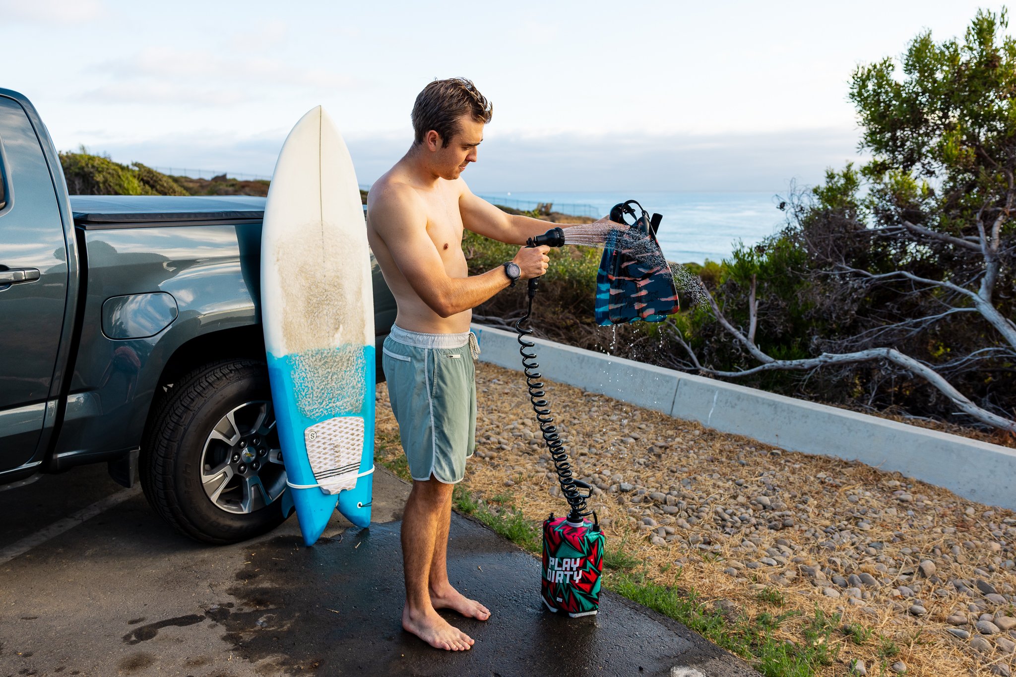 A young man wearing swim trunks rinses off his swimsuit with a garden hose next to a pile of surfboards and a pickup truck, with a coastal landscape in the background.