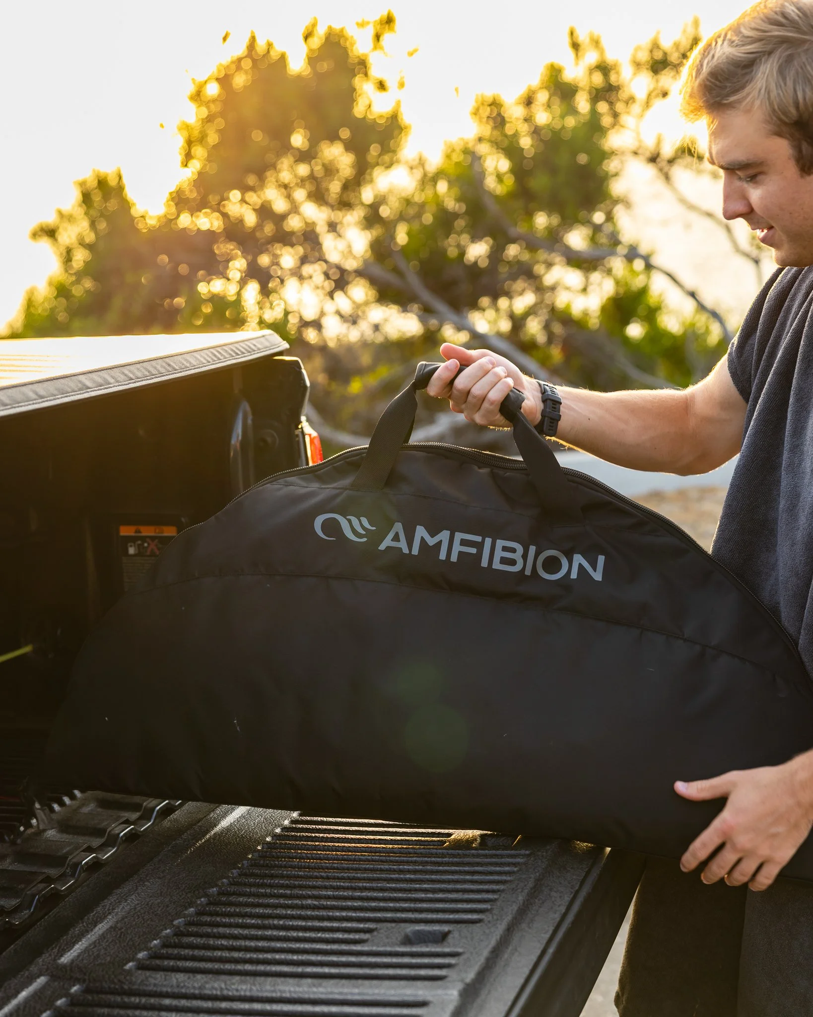 A man loading a black bag labeled 'AMFIBION' into the back of a pickup truck, outdoors during sunset with trees in the background.