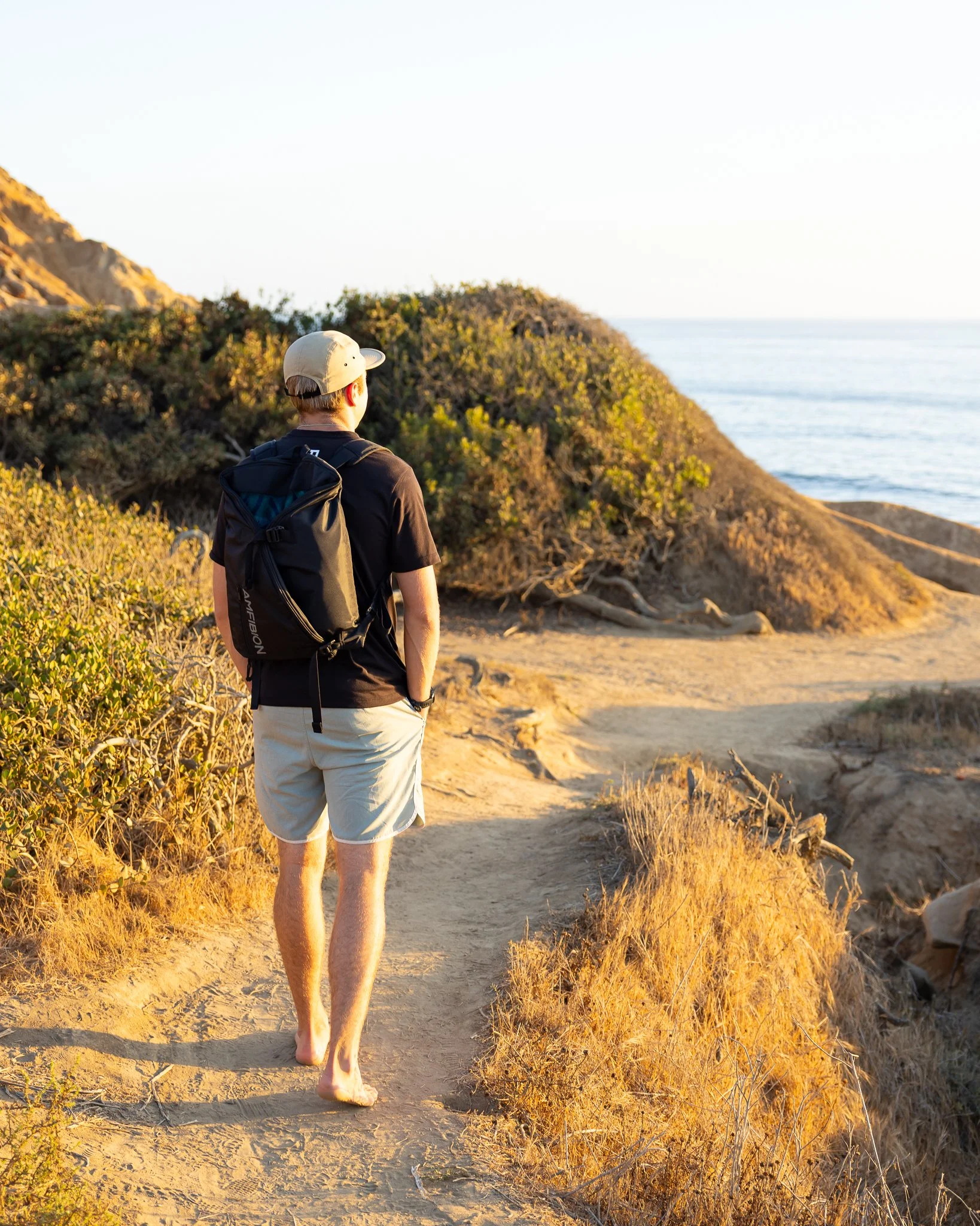 A man walking barefoot on a dirt trail along a rocky coastal landscape with shrubs and the ocean in the background during sunset.