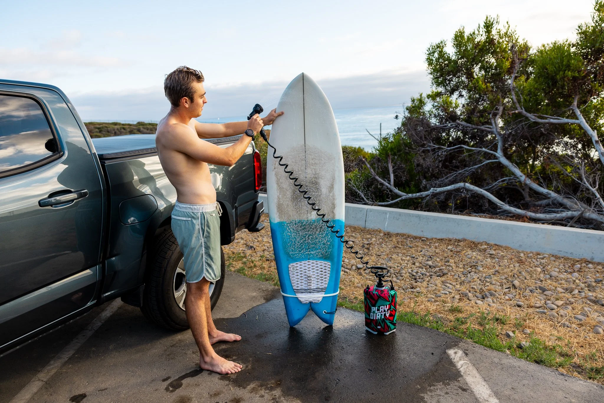 A man in swim trunks is drying off after surfing, standing next to a surfboard and a backpack spray bottle, in a parking lot near the beach with trees and the ocean in the background.