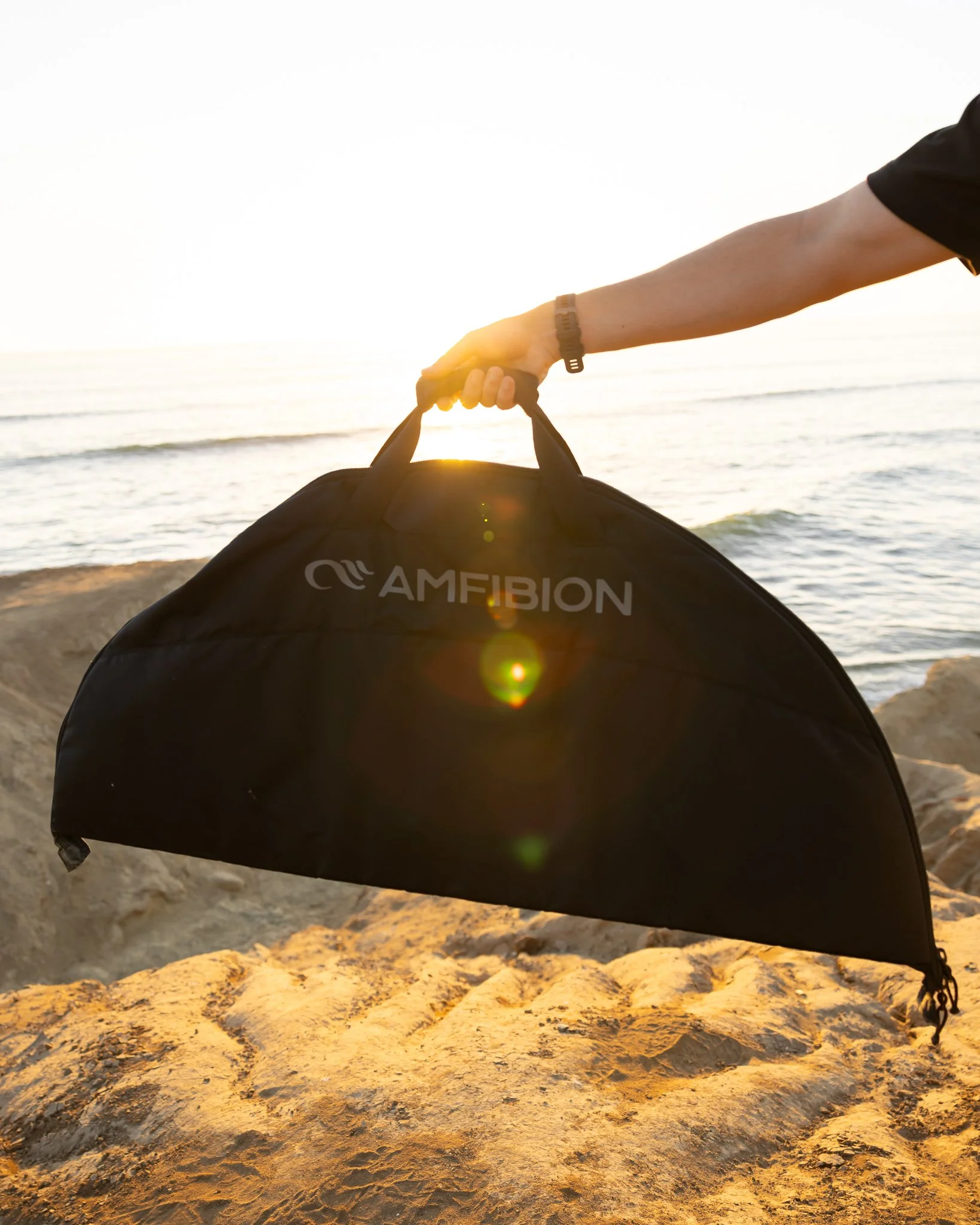 Person holding a black sleek carrying case labeled 'AMFIBION' on a sandy beach at sunset by the ocean.