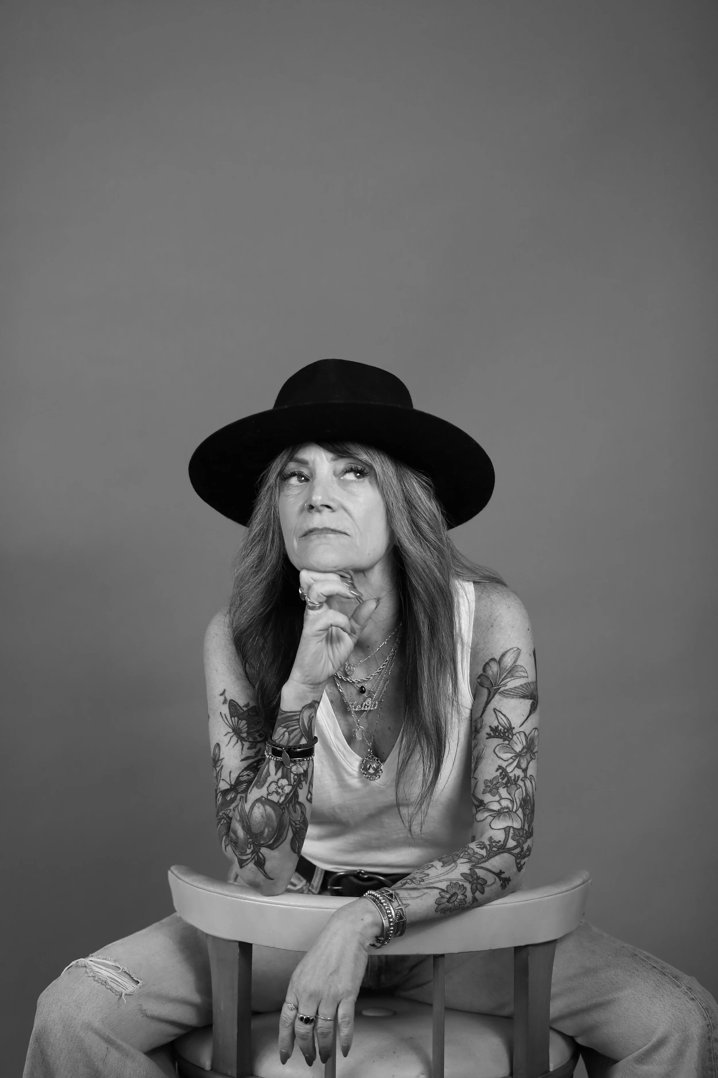 Black and white photo of a woman with long hair, tattoos on her arms, wearing a black hat, tank top, and multiple jewelry, sitting on a high chair with her legs apart, resting her elbows on her knees and her chin on her hand, against a plain backgrou