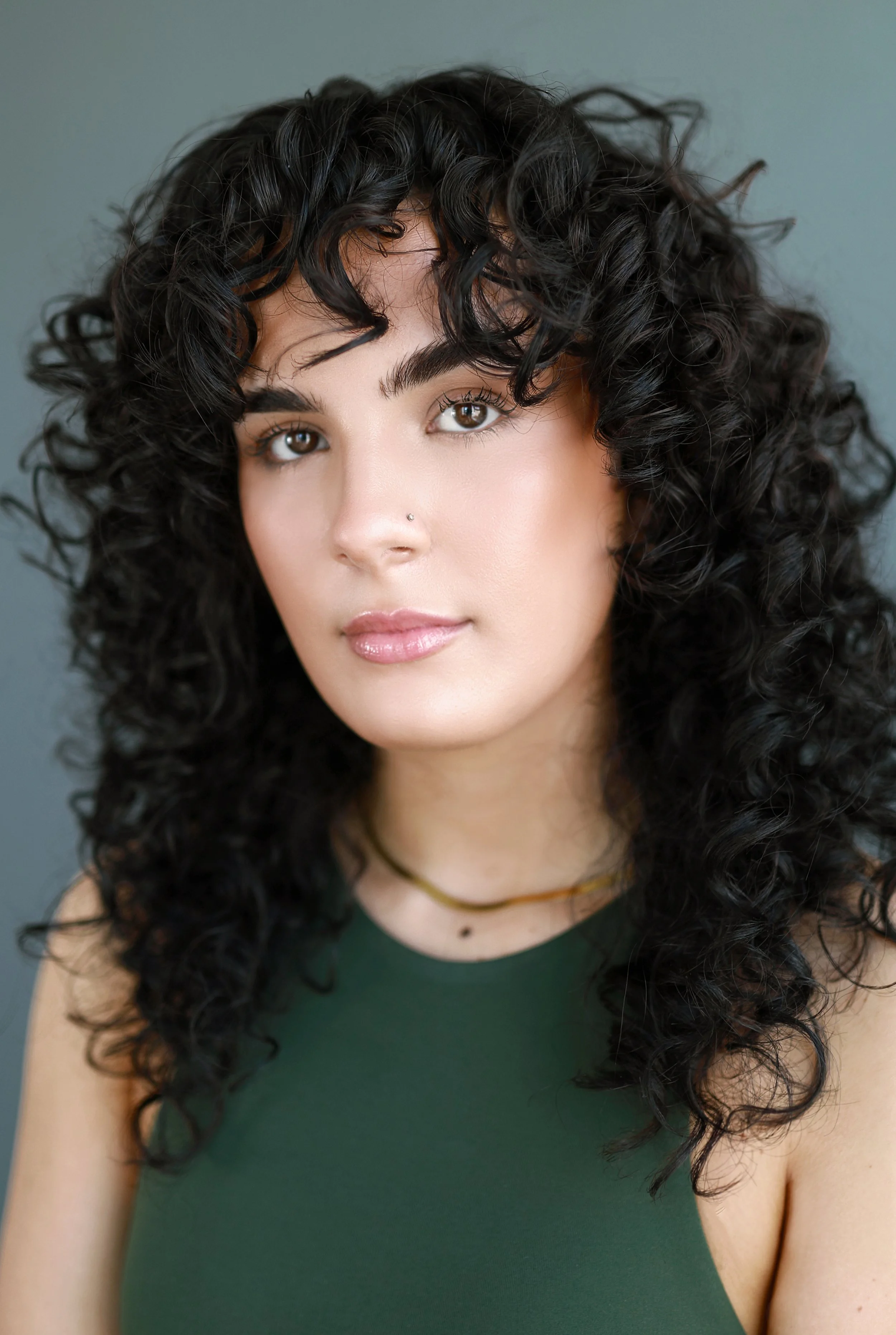 Close-up of a young woman with dark, curly hair, wearing a green sleeveless top, with natural makeup and a small nose piercing.