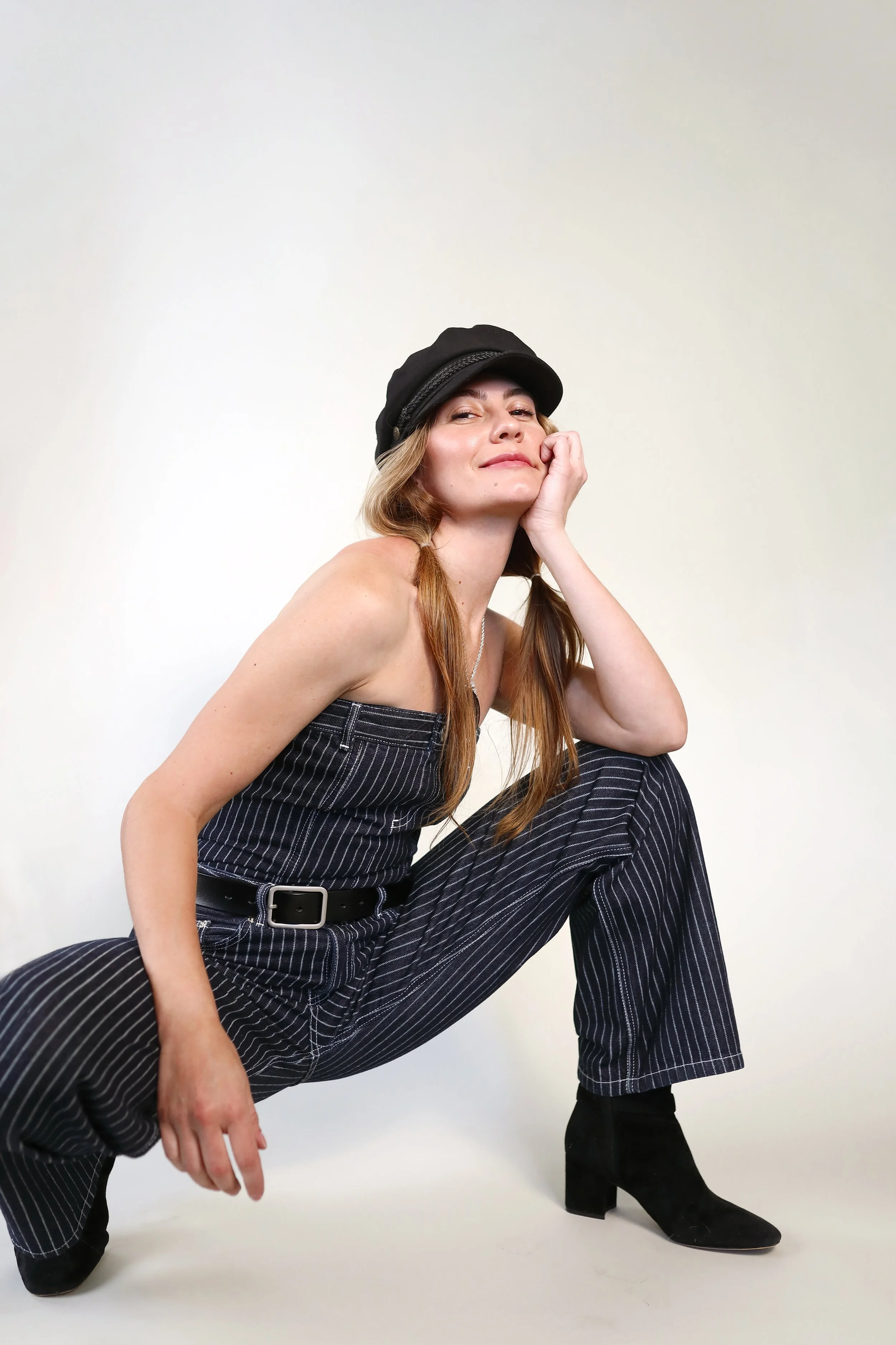 A woman with long hair, wearing a black cap and pinstripe pants, is sitting with one knee up, resting her head on her hand, and smiling at the camera. She is also wearing black heeled boots.