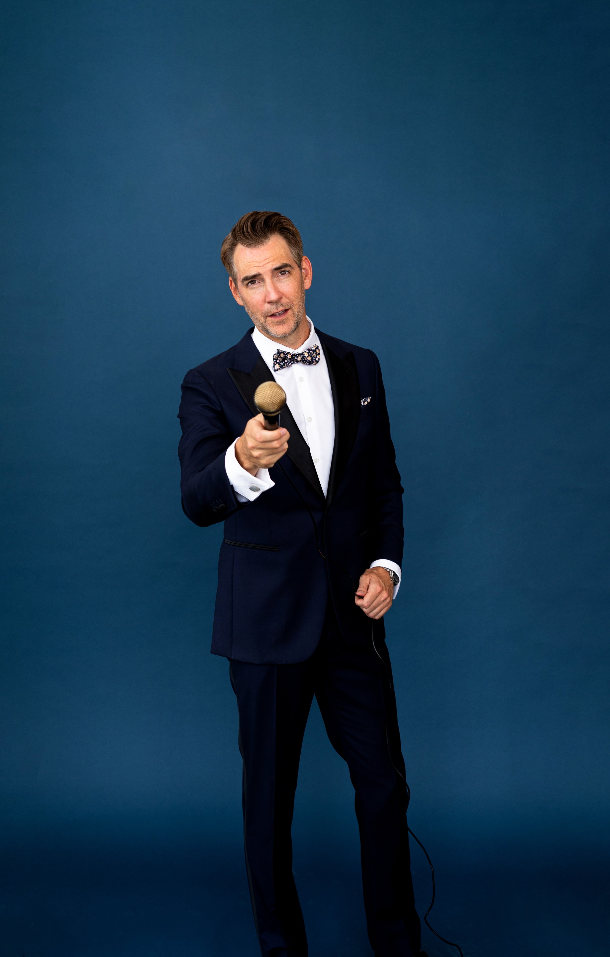 A man in a dark tuxedo with a bow tie holding a microphone, standing against a plain blue background.