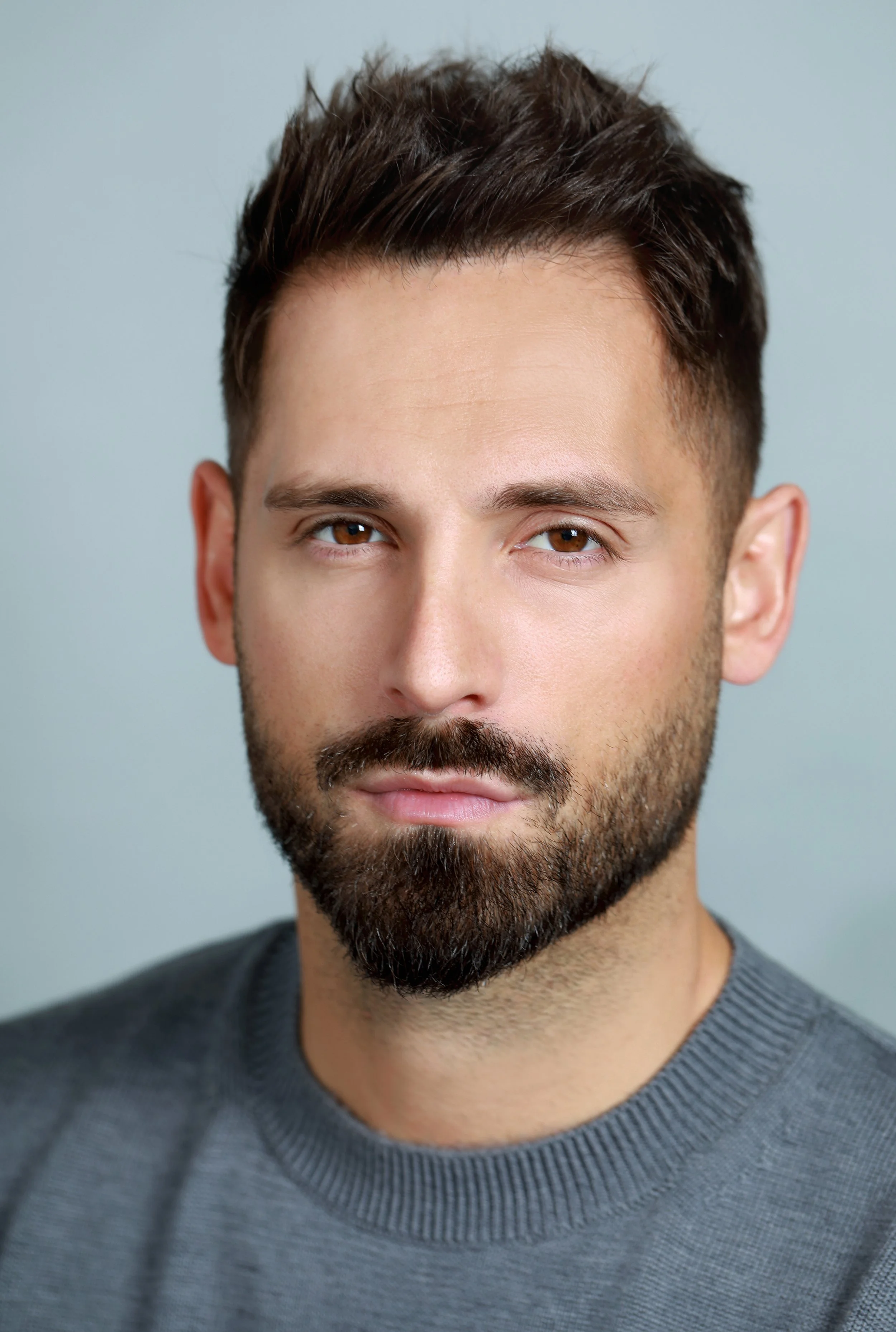 Close-up of a man's face with brown eyes, dark hair, and a beard, wearing a gray sweater.