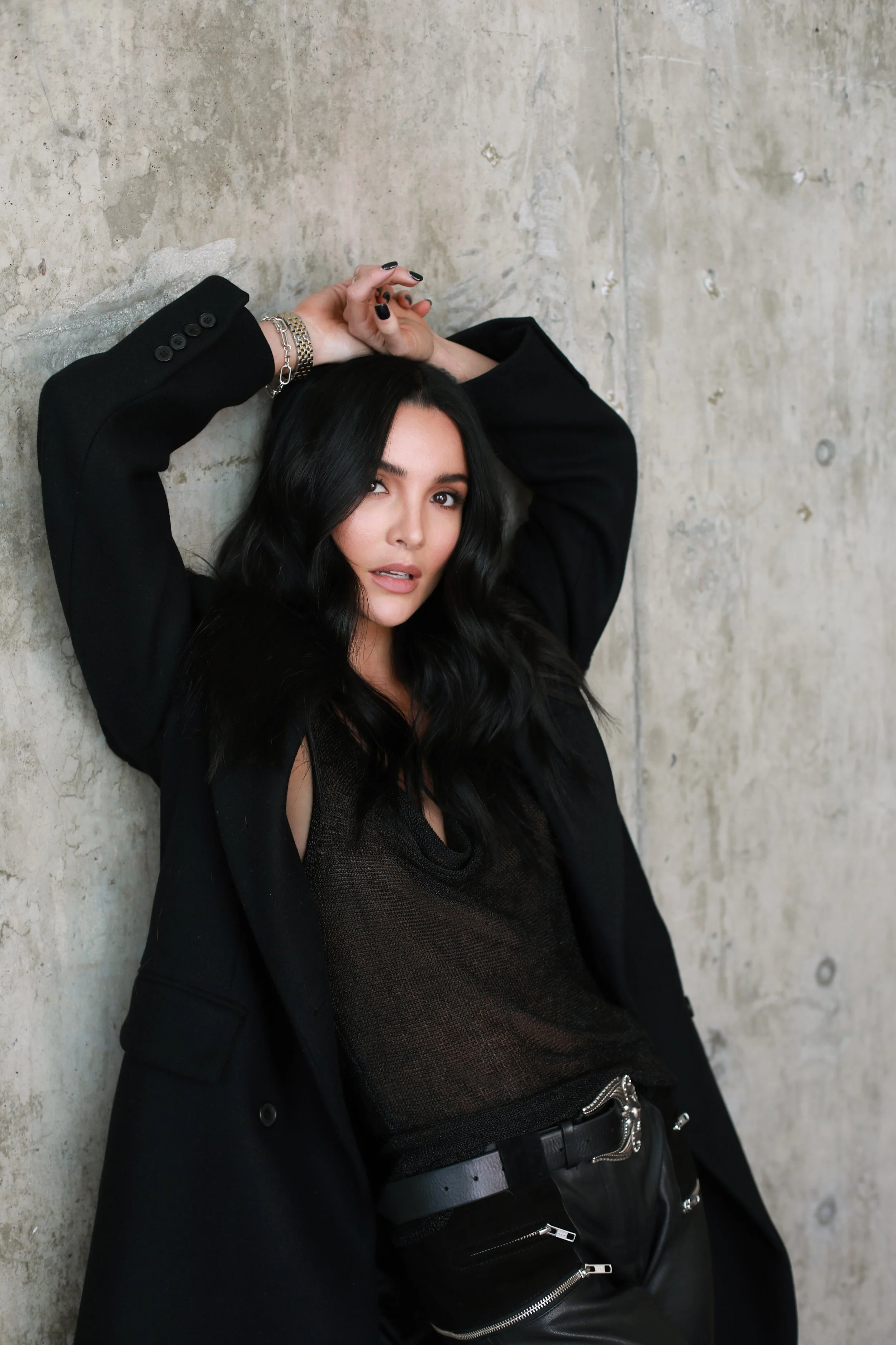 A woman with long black hair and light skin gender expression, wearing a black blazer, gray mesh top, and black leather pants, is leaning against a concrete wall. She has silver jewelry on her wrist and belt, black nail polish, and is looking at the camera with a slightly open mouth.