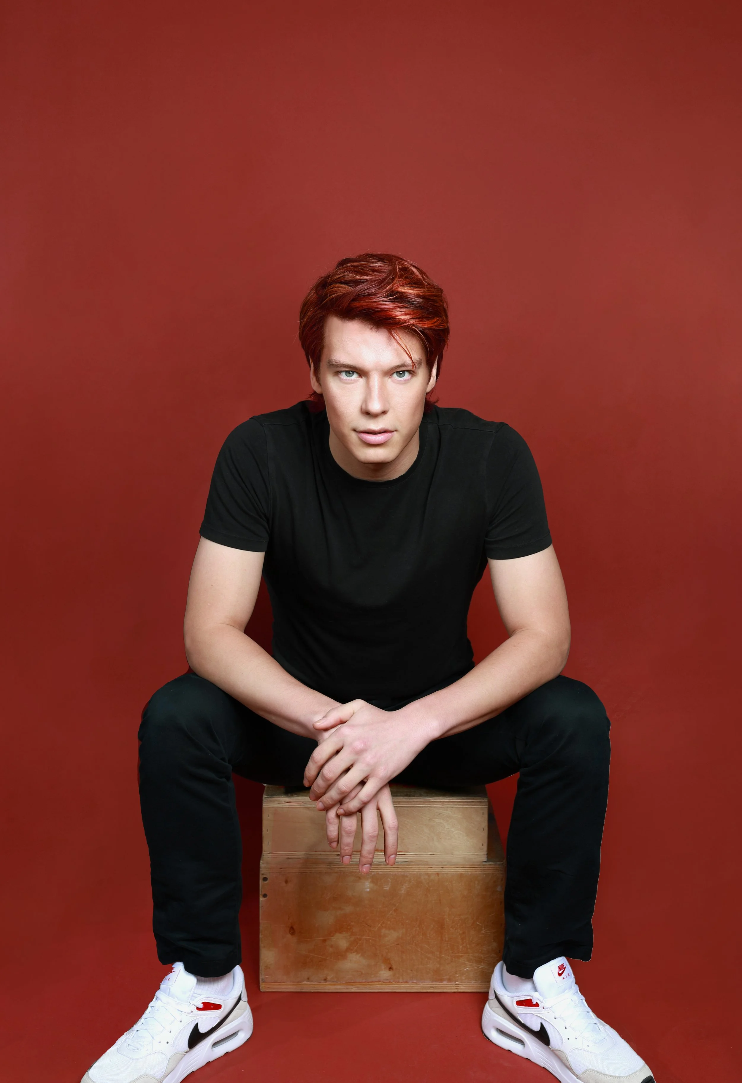 Young man with red hair, wearing a black t-shirt and black pants, sitting on a wooden box against a red background, with his hands clasped in front of him and wearing white Nike sneakers.