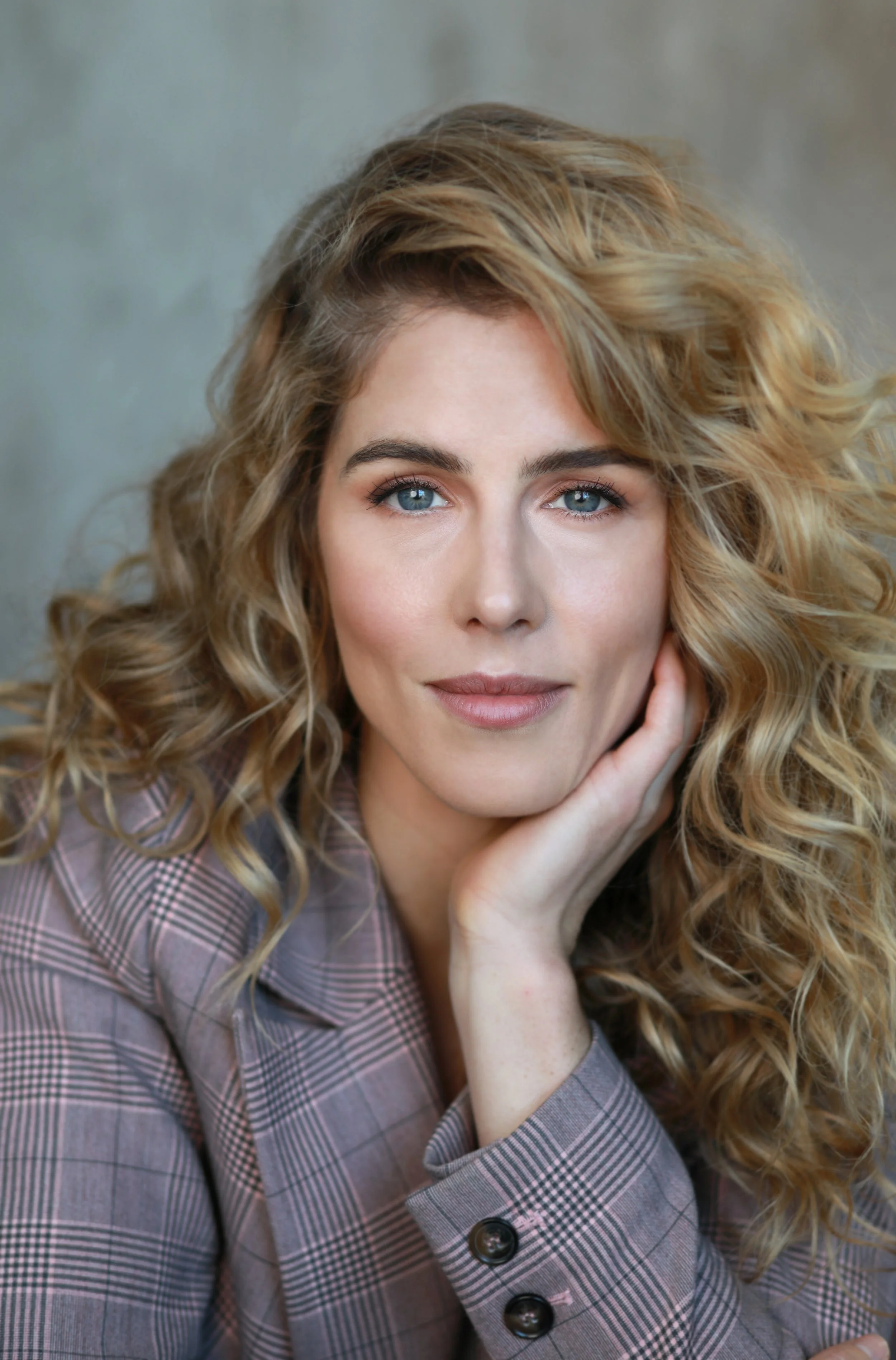 A woman with long, curly blonde hair and blue eyes, wearing a plaid blazer with large buttons, posing with her hand resting on her face.