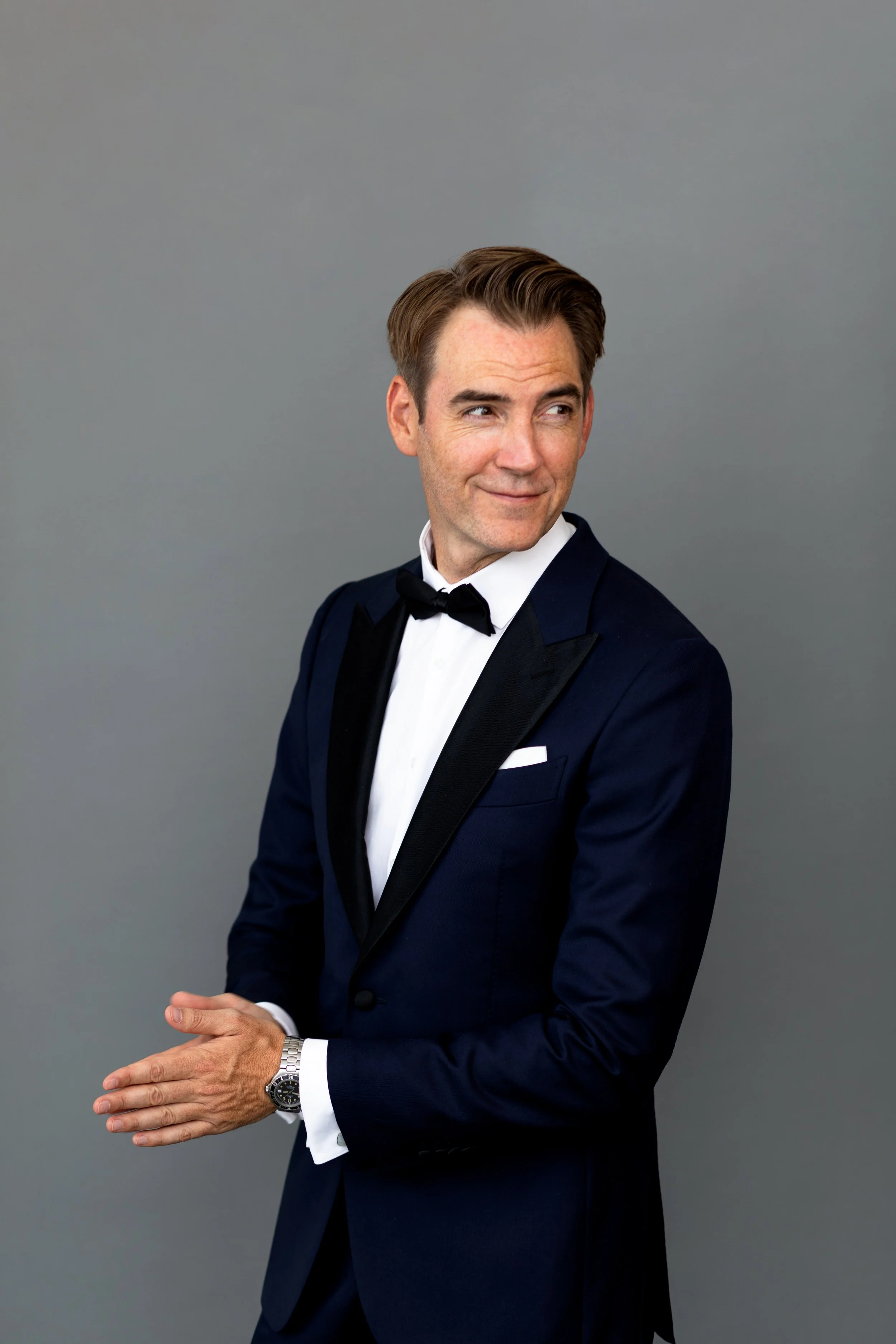 A man with light skin and brown hair in a slicked-back hairstyle, wearing a navy blue tuxedo with a black satin lapel, a white dress shirt, and a black bow tie. He is standing against a plain gray background, with his hands clasped in front of him, a