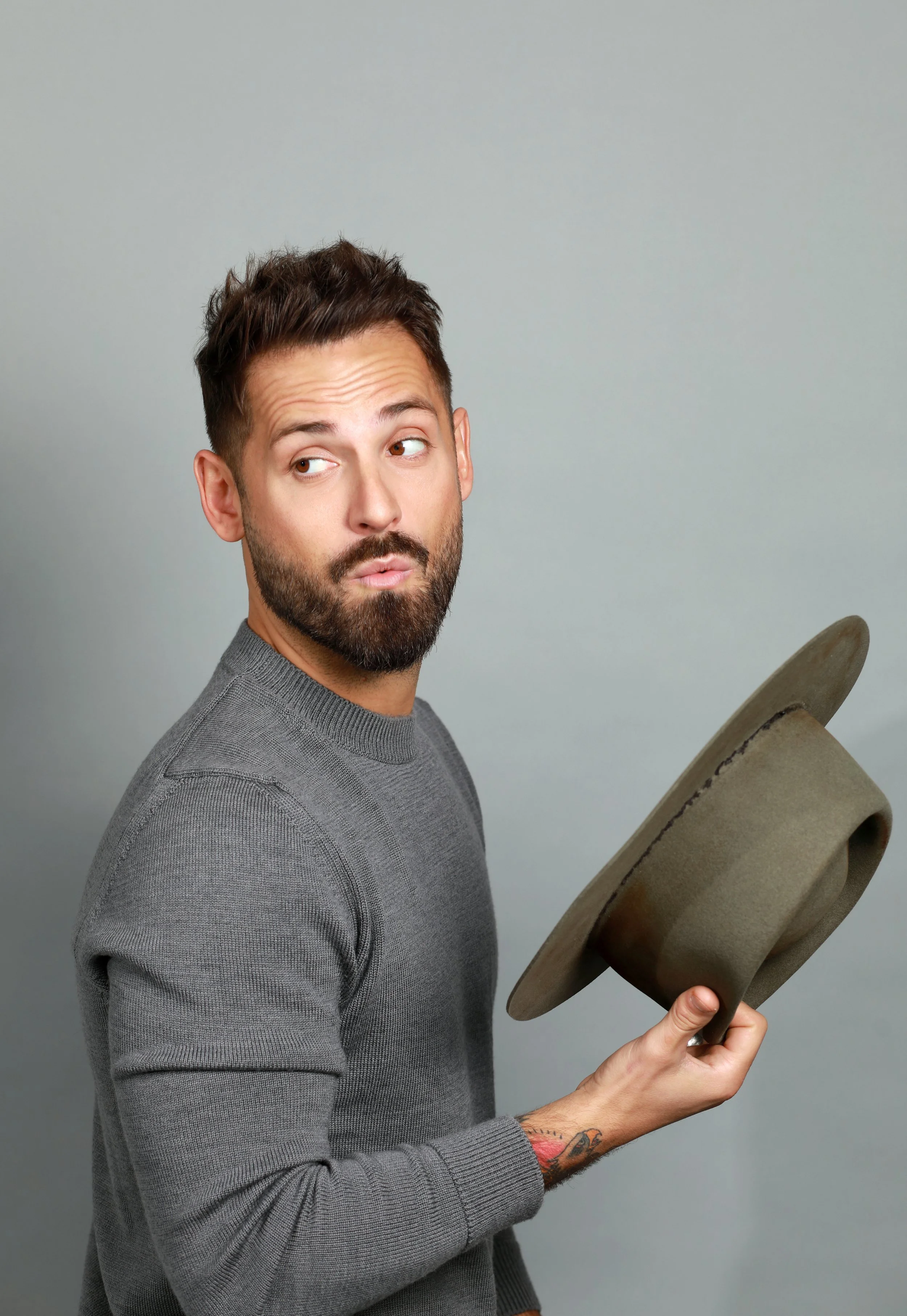 A man with a beard and short brown hair looking to the side, wearing a gray sweater, holding a brown fedora hat in his right hand, with a tattoo on his left wrist, against a plain gray background.