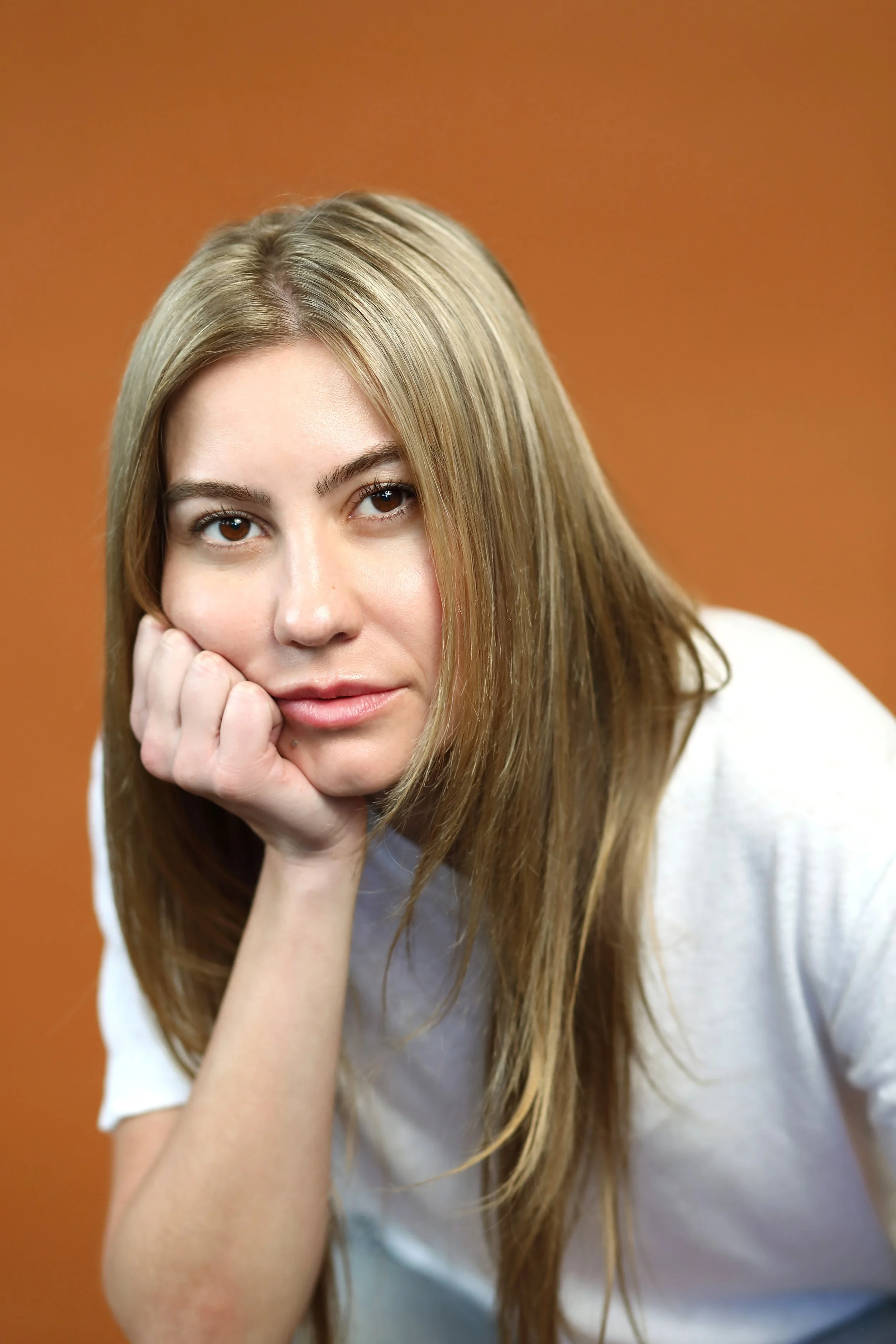 Close-up of a young woman with long blonde hair, resting her chin on her hand, looking at the camera against a brown background.