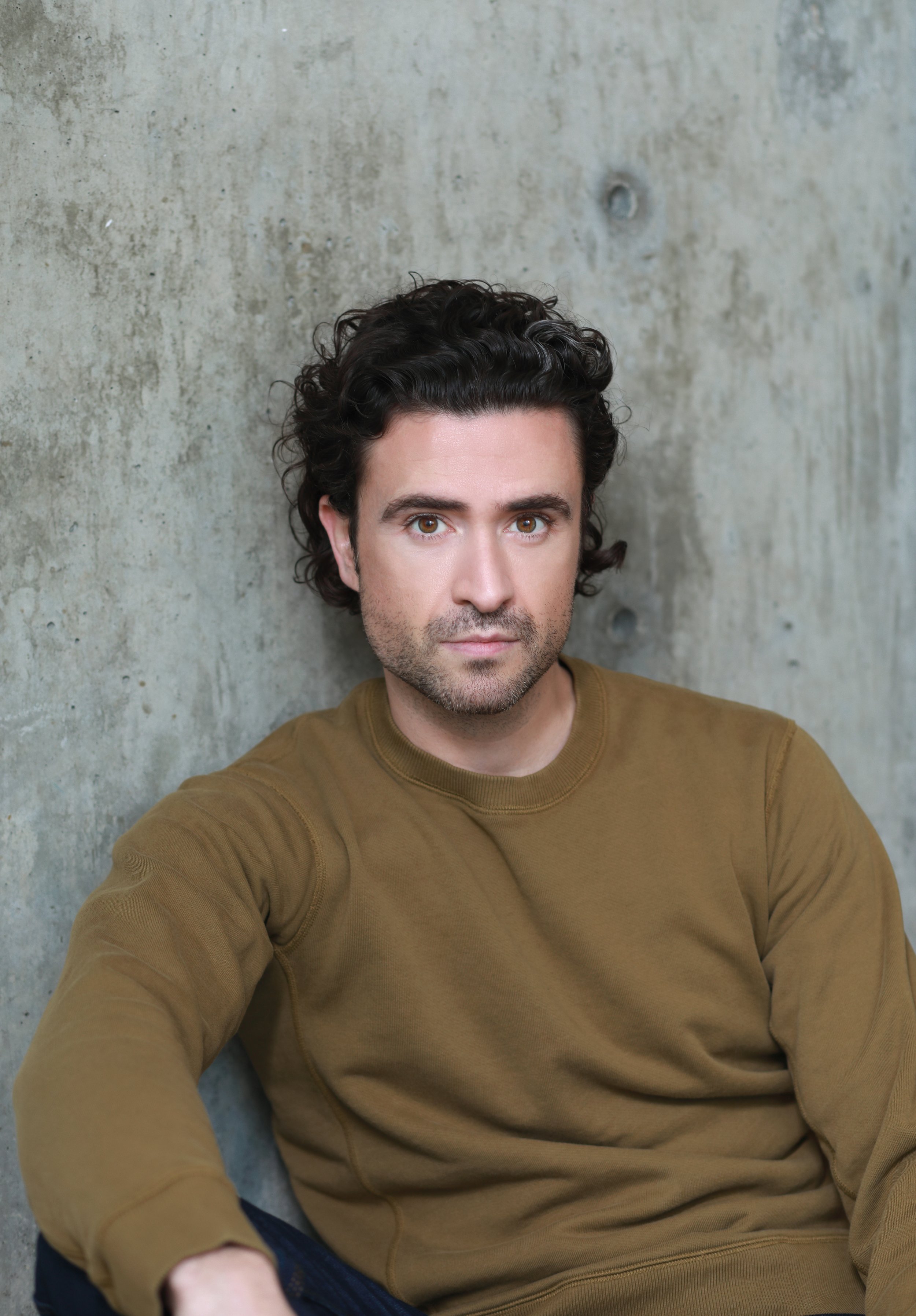 A man with dark curly hair and brown eyes wearing a brown long sleeve shirt, taking a selfie against a concrete wall.