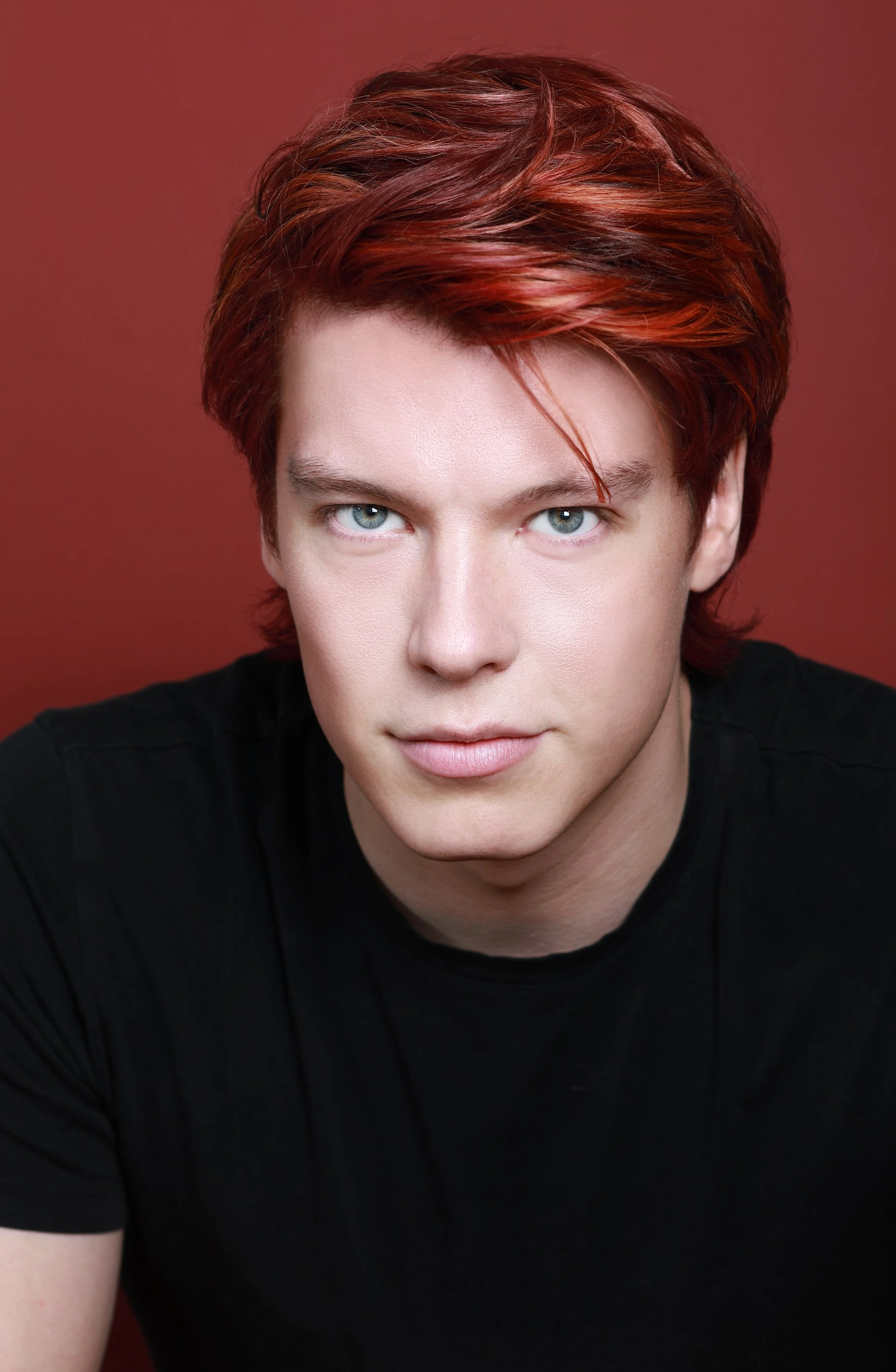 A young person with short, red hair and blue eyes, wearing a black shirt, against a red background.