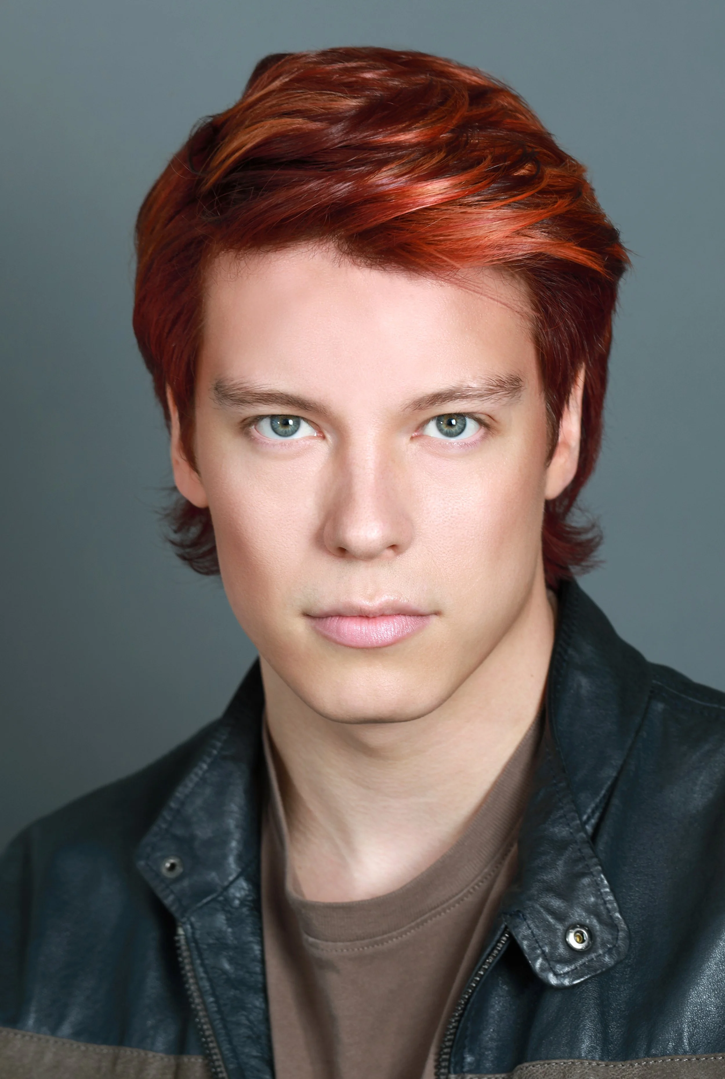 Close-up of a person with short red hair and blue eyes, wearing a black leather jacket and a brown shirt, against a gray background.