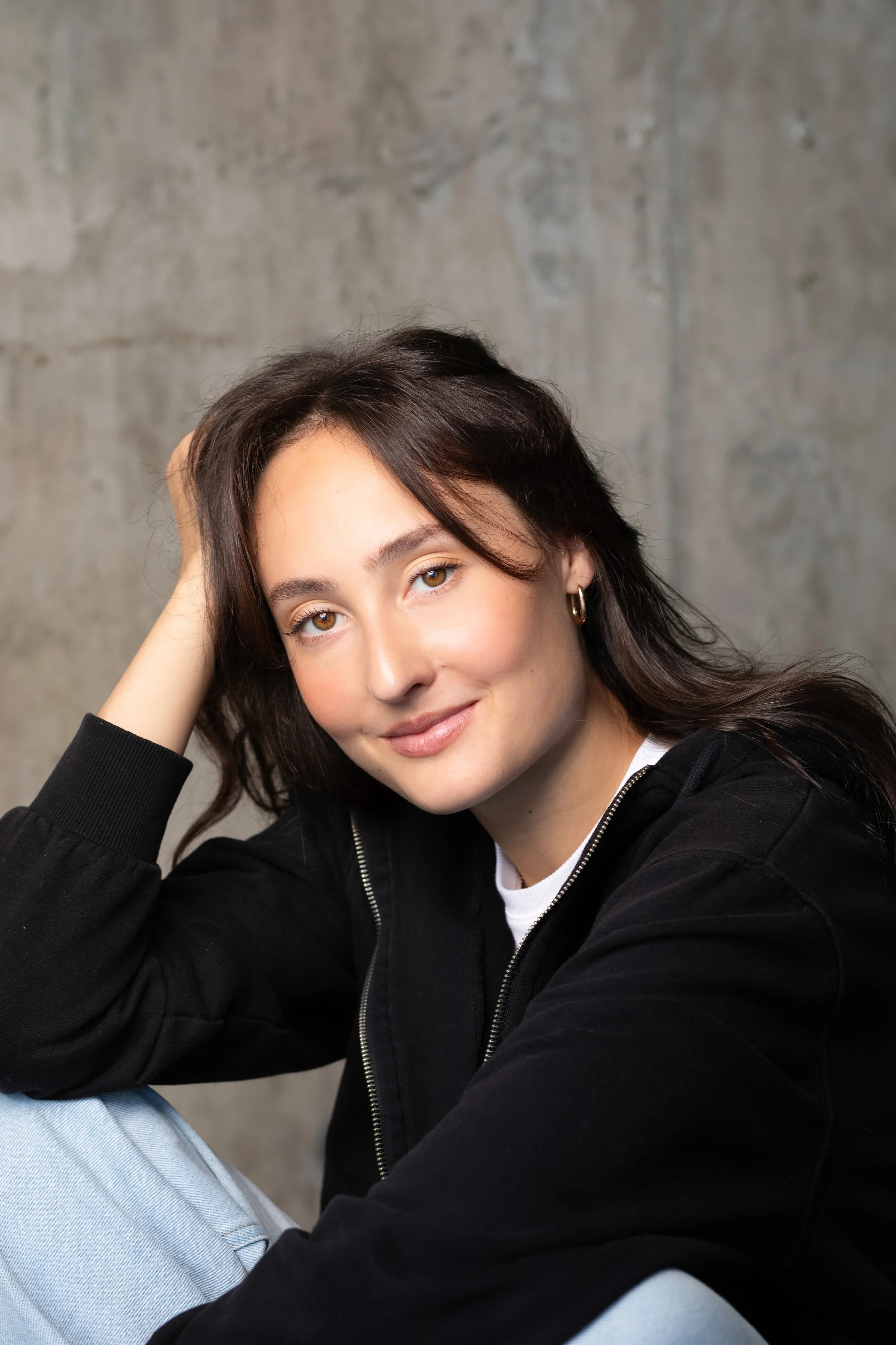 A young woman with dark hair and brown eyes smiling, wearing hoop earrings and a black zip-up jacket, sitting against a grey concrete wall.