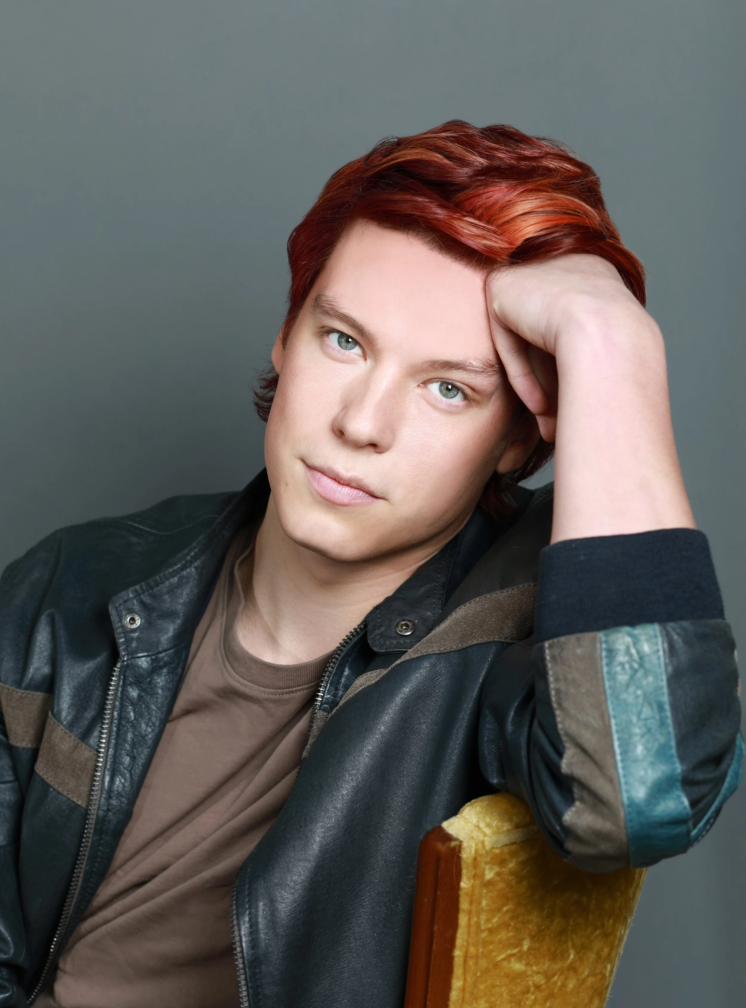Young man with reddish hair and blue eyes sitting on a yellow chair, resting his head on his hand, wearing a black leather jacket and a brown shirt, against a gray background.