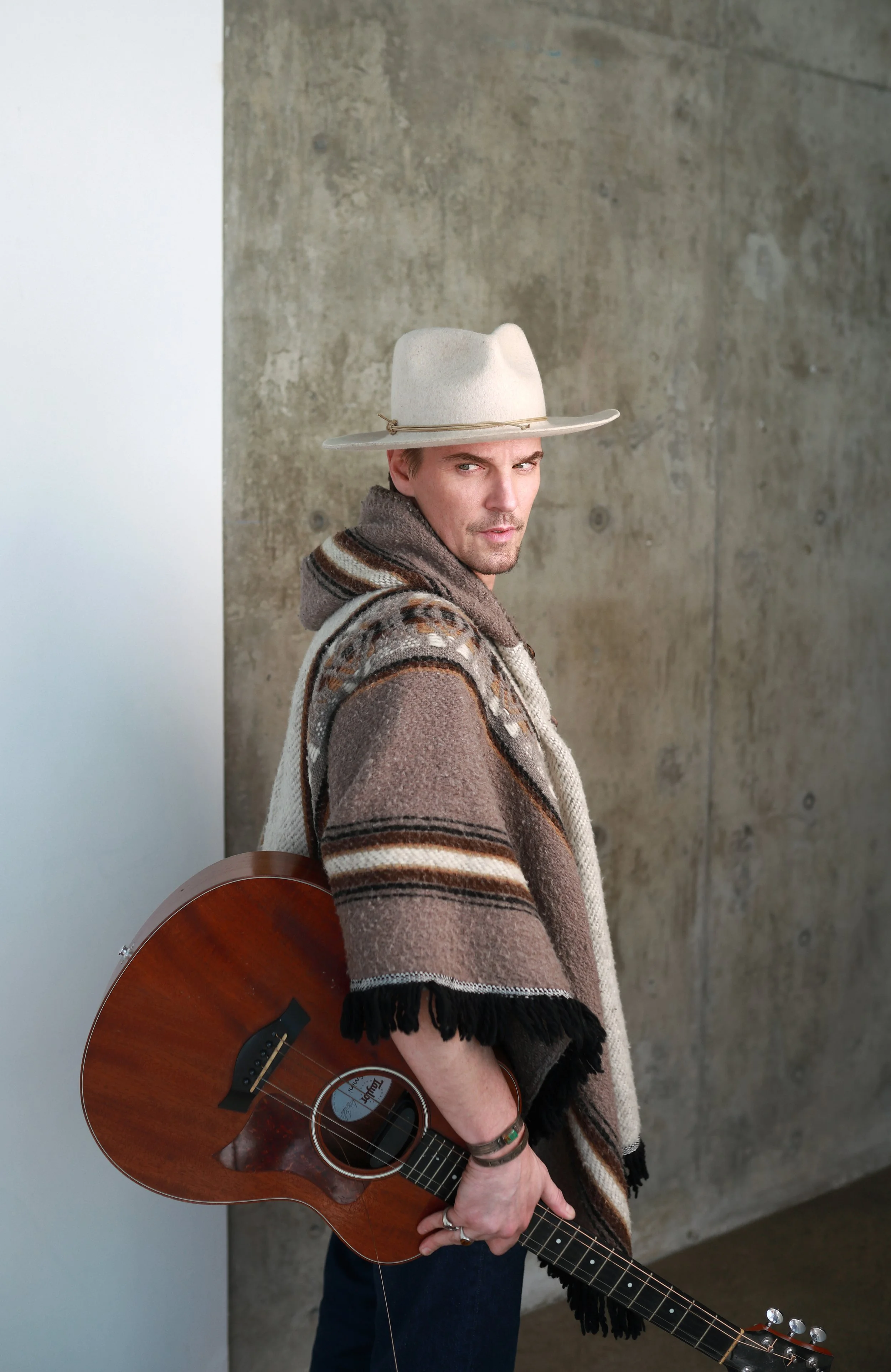 A man in a wide-brimmed hat and patterned sweater holding an acoustic guitar next to a concrete wall.