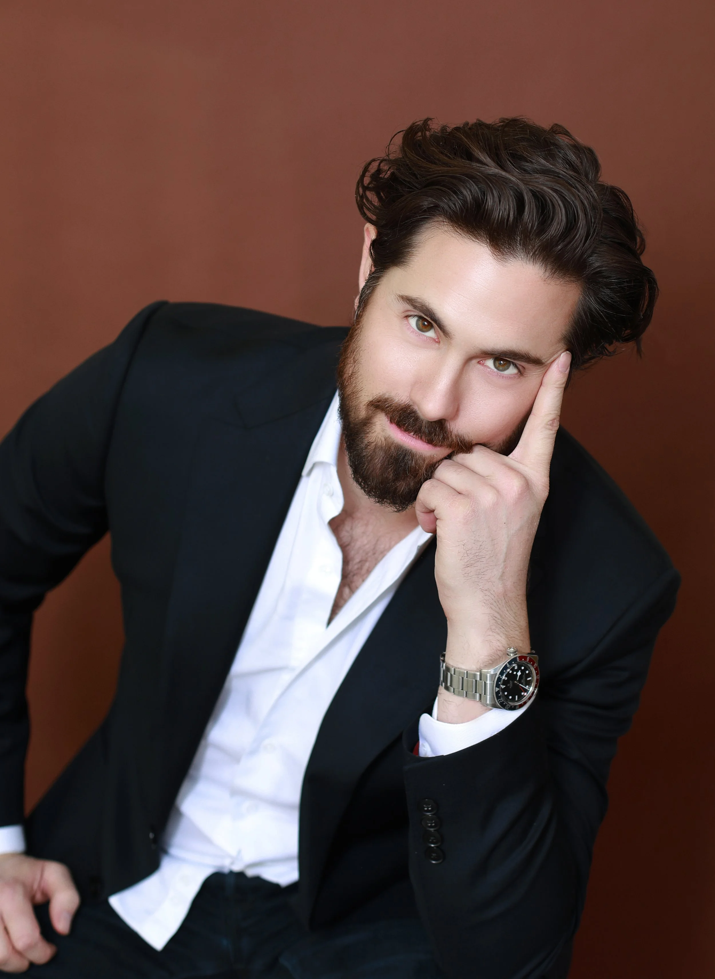 A man with brown hair and beard wearing a dark blazer, white shirt, and silver watch, casually leaning with his hand on his face against a plain brown background.