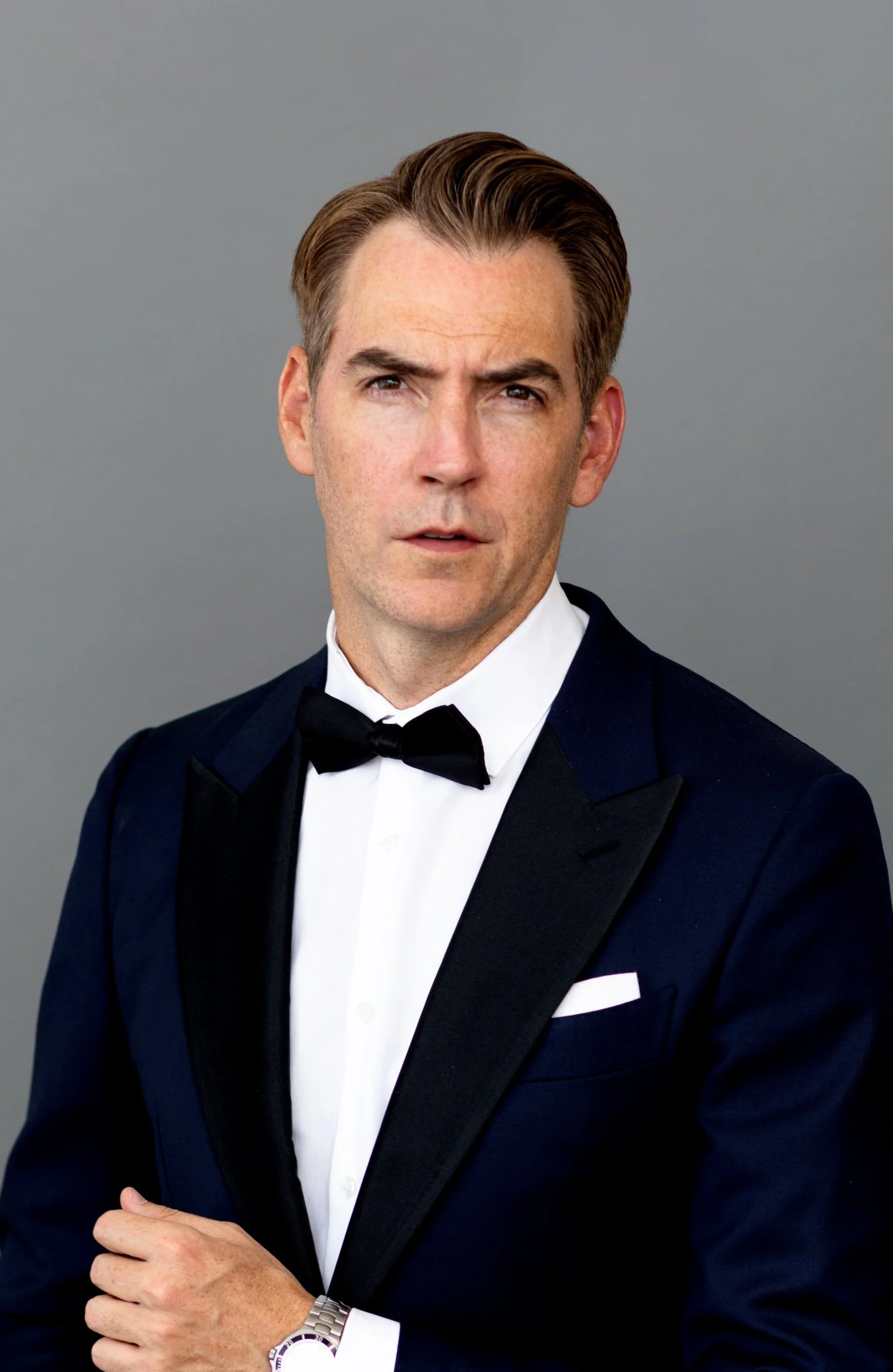 A man in a tuxedo with a black bow tie, white shirt, and silver watch posing against a plain gray background.