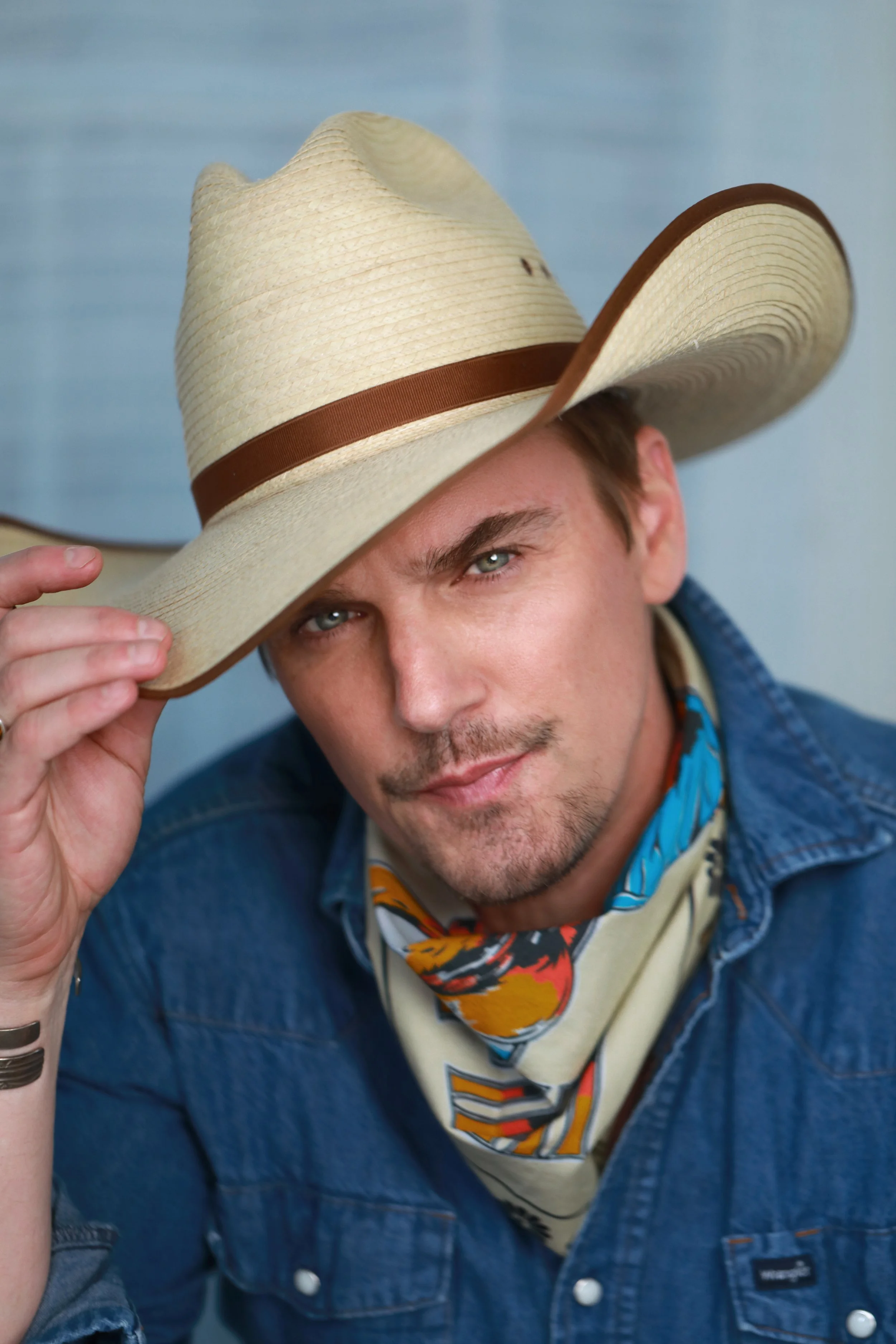 A young man with light skin and piercing blue eyes wearing a large straw cowboy hat with a brown band, a colorful bandana, and a denim jacket, holding the brim of his hat with a slight smile, against a simple blue background.