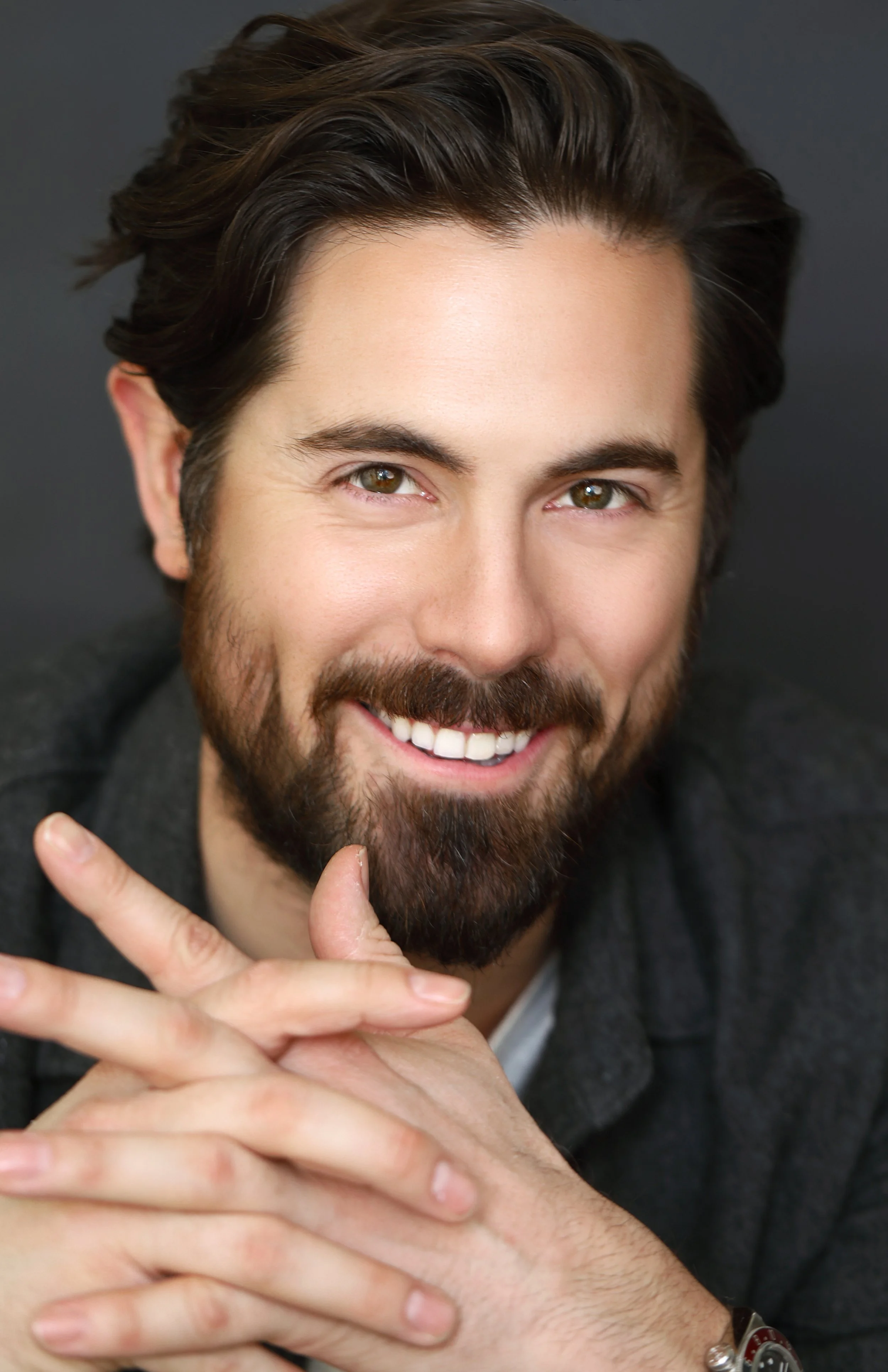 Close-up of smiling man with dark hair and beard, showing brown eyes, wearing a dark blazer, holding his hands in front of him.