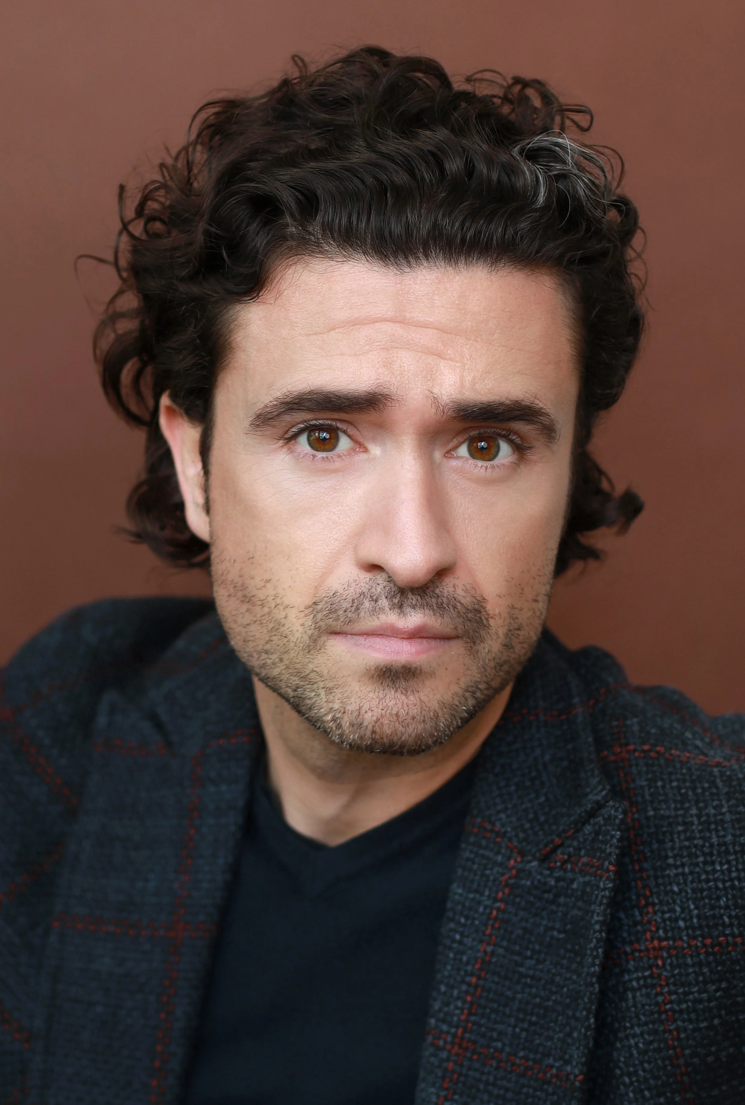 A young man with dark, curly hair and hazel eyes taking a selfie against a plain, brown background. He has light skin and facial hair and is wearing a dark shirt with a plaid, dark-colored blazer.