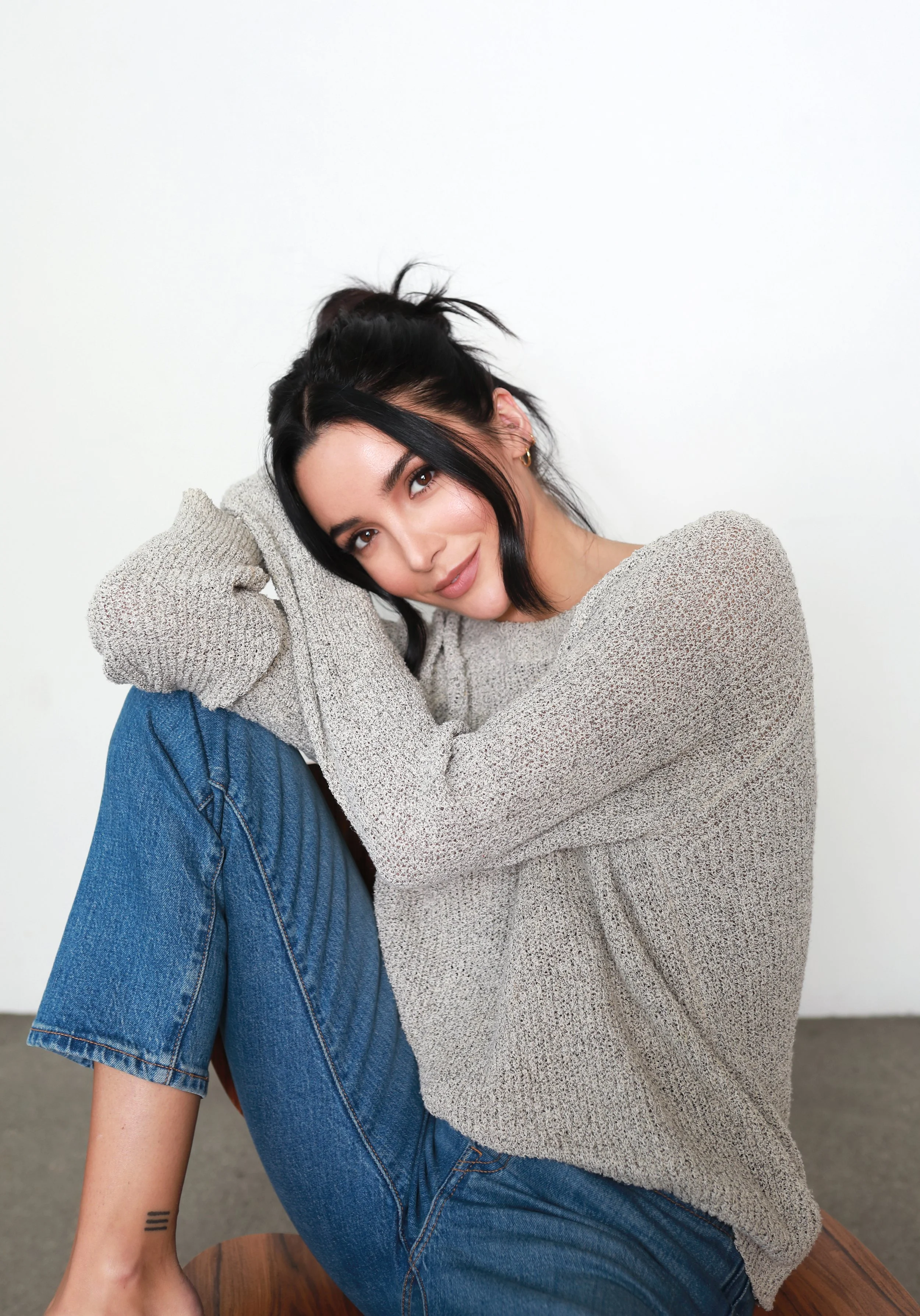 Young woman with black hair in a messy bun and a small tattoo on her wrist, wearing a beige sweater and blue jeans, sitting on a wooden surface against a plain white wall.